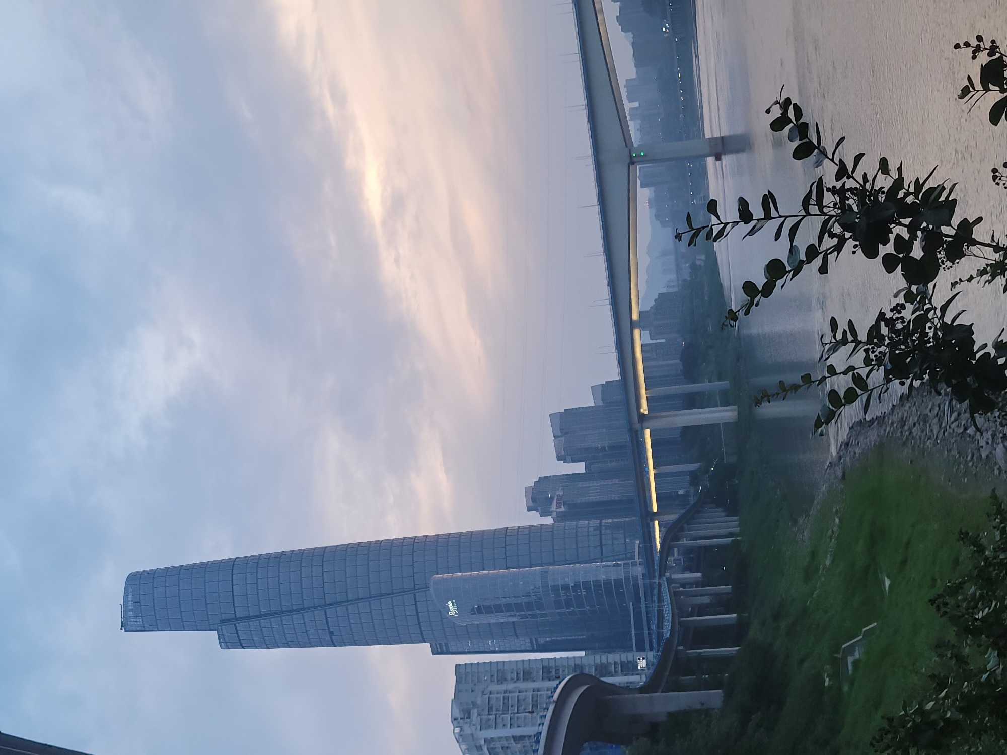 The image captures a dusk scene in Yuzhong District, China. A tall, modern skyscraper with a cylindrical section dominates the left side. Behind it, other buildings are visible. A monorail track curves around the base of the buildings. A bridge spans a wide body of water, with the city skyline visible in the background. Lush vegetation is present on the right side of the frame, obscuring the view of the water. The sky is overcast with clouds reflecting the setting sun.