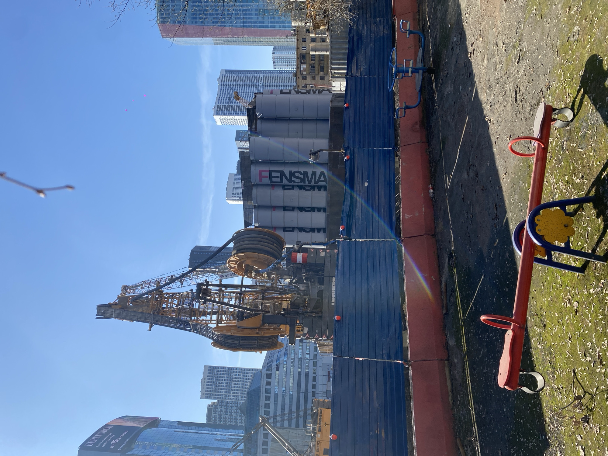 The image shows a construction site in Khimki, Russia. A large yellow crane dominates the scene, positioned in front of cylindrical silos bearing the "FENSMA" logo. A blue construction fence separates the site from a public area, possibly a playground, indicated by the presence of a seesaw. The background features numerous tall buildings, typical of an urban environment. The sky is clear and blue.
