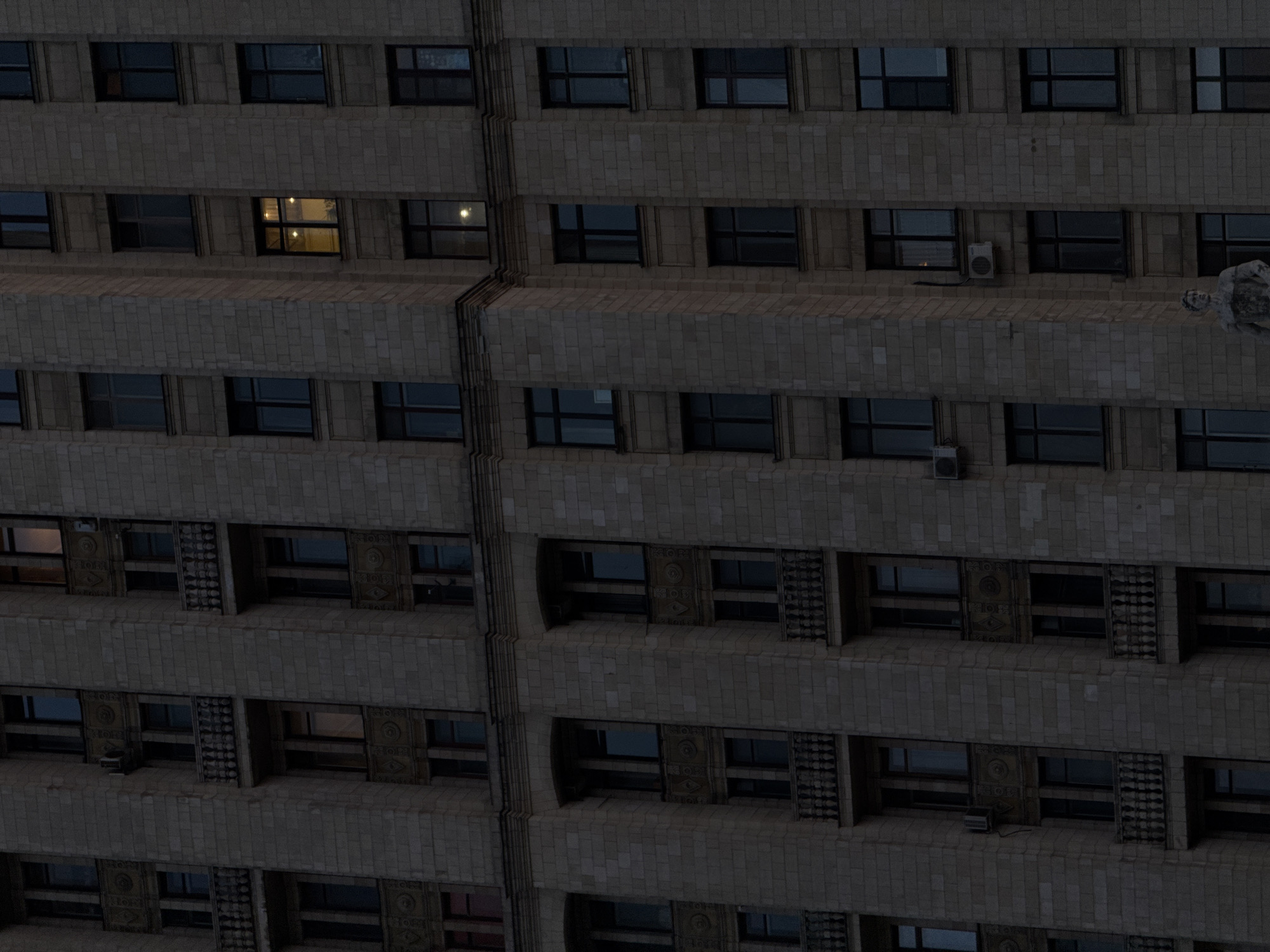 A low-angle, medium-shot photograph depicts a multi-story building at dusk. The building, located in Moscow, Russia, is made of beige bricks and features rows of windows. Most of the windows are dark, reflecting the ambient light, while one window on the third story glows with interior light. A few air conditioning units are attached to the building's exterior. The building appears to be of a Soviet-era architectural style.