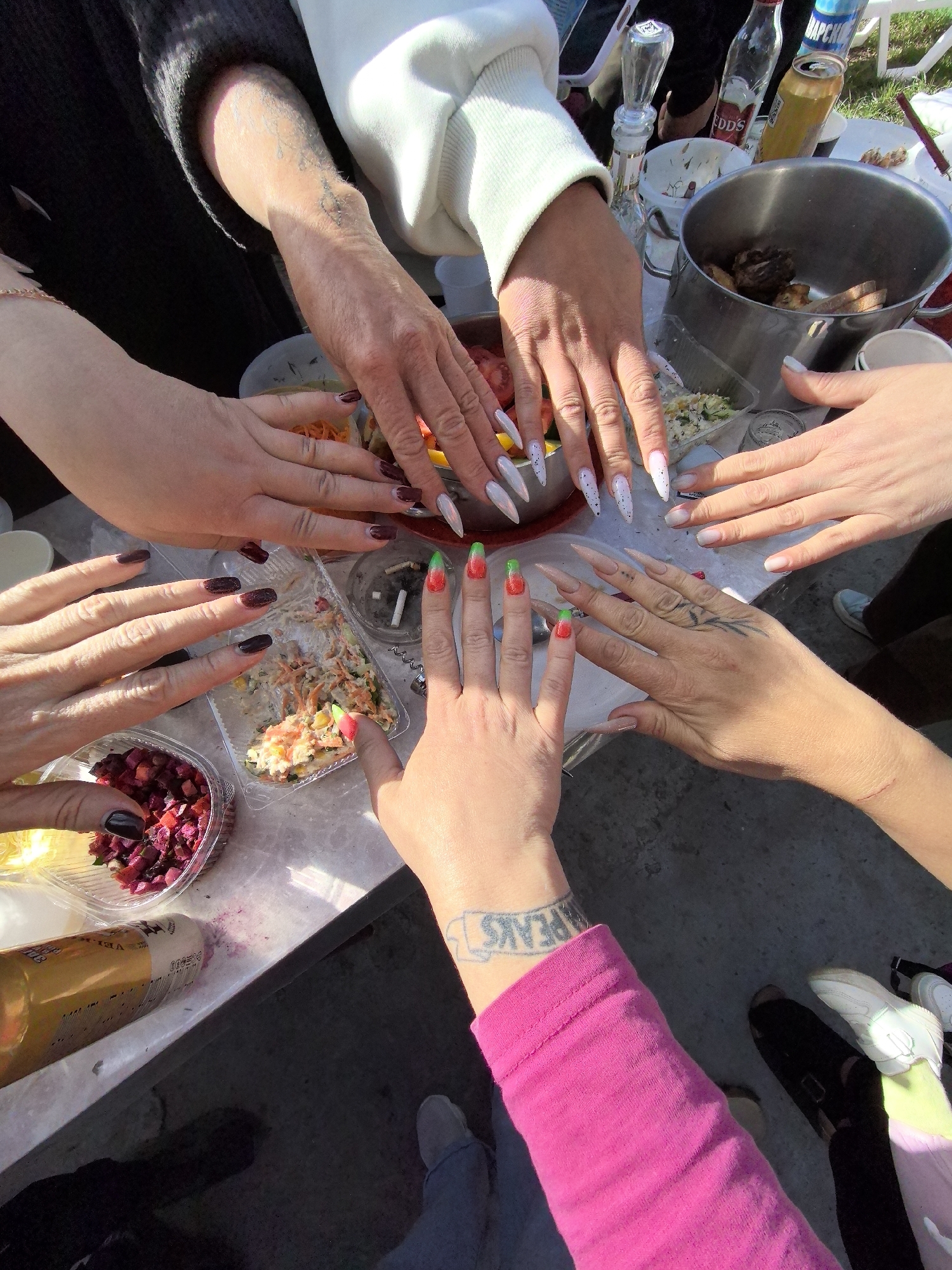 A top-down shot shows a group of people gathered around a table filled with food containers and beverages. The table appears to be outdoors, possibly in a park or garden setting. Various hands reach toward the table, some adorned with painted nails. A pot of meat sits among the food containers. Beverages include bottles of what appears to be alcohol, as well as canned drinks. A woman in a pink long-sleeved shirt and a tattoo on her wrist is standing near a pair of feet wearing shoes. The location is not identifiable based on the information provided.