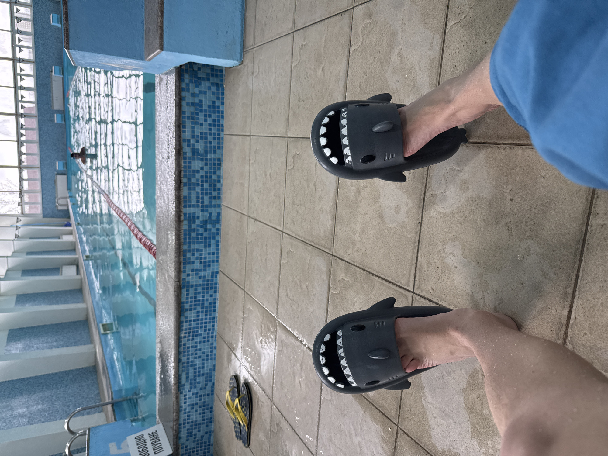The image shows a person standing on a tiled floor next to a swimming pool. The person is wearing blue shark-shaped slippers. In the background, there is a swimming pool with a person swimming in it. There is a red rope dividing the pool. Yellow and black flip-flops are visible on the floor near the pool's edge. The location is likely a swimming pool area, potentially in Plovdiv, Bulgaria, but there is insufficient information in the picture to confirm the city of Plovdiv.