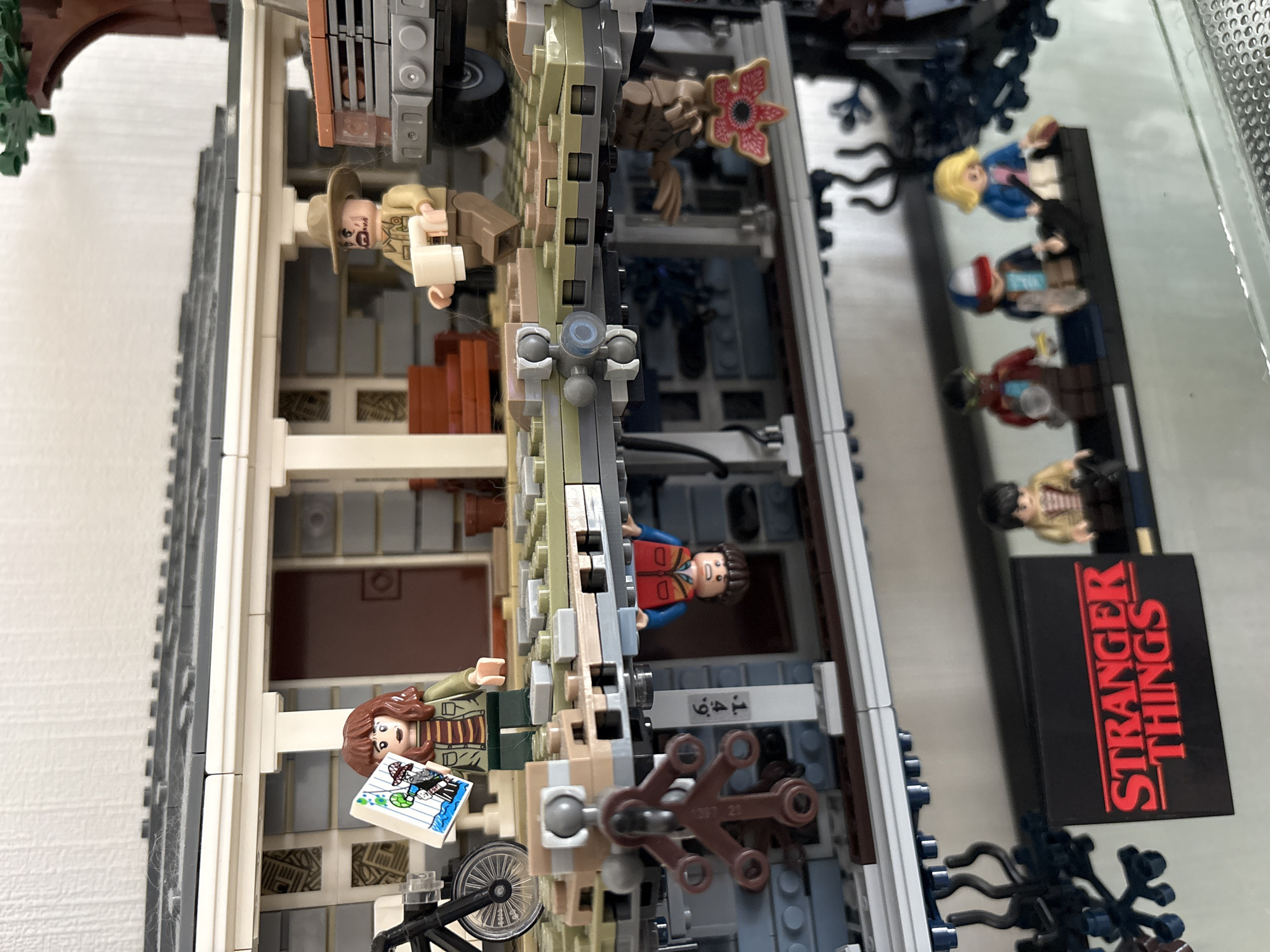 This image displays a Lego set of the Byers' house from the television series "Stranger Things", positioned sideways. The set is two-sided, depicting the house in the normal world on one side and the Upside Down on the other. Several Lego minifigures, including Eleven, Mike, Dustin, Lucas, and Will, are placed beside the set on a reflective surface. The "Stranger Things" logo is also visible next to the minifigures. The overall setting appears to be indoors.