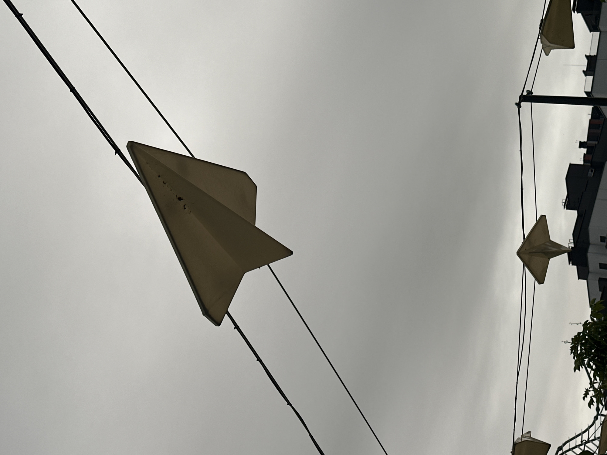 An off-white, worn, paper airplane-shaped ornament is prominently displayed against an overcast, light grey sky in Бараўлянскі сельскі Савет, Belarus. It is suspended in the upper half of the frame by two dark, diagonal wires. Multiple dark electrical or support wires traverse the scene, with additional similar paper airplane ornaments partially visible along a lower horizontal set of wires. Below these wires, green foliage partially obstructs the view of dark-roofed buildings with lighter facades in the background. A black vertical pole is visible on the lower right, anchoring some of the wires.