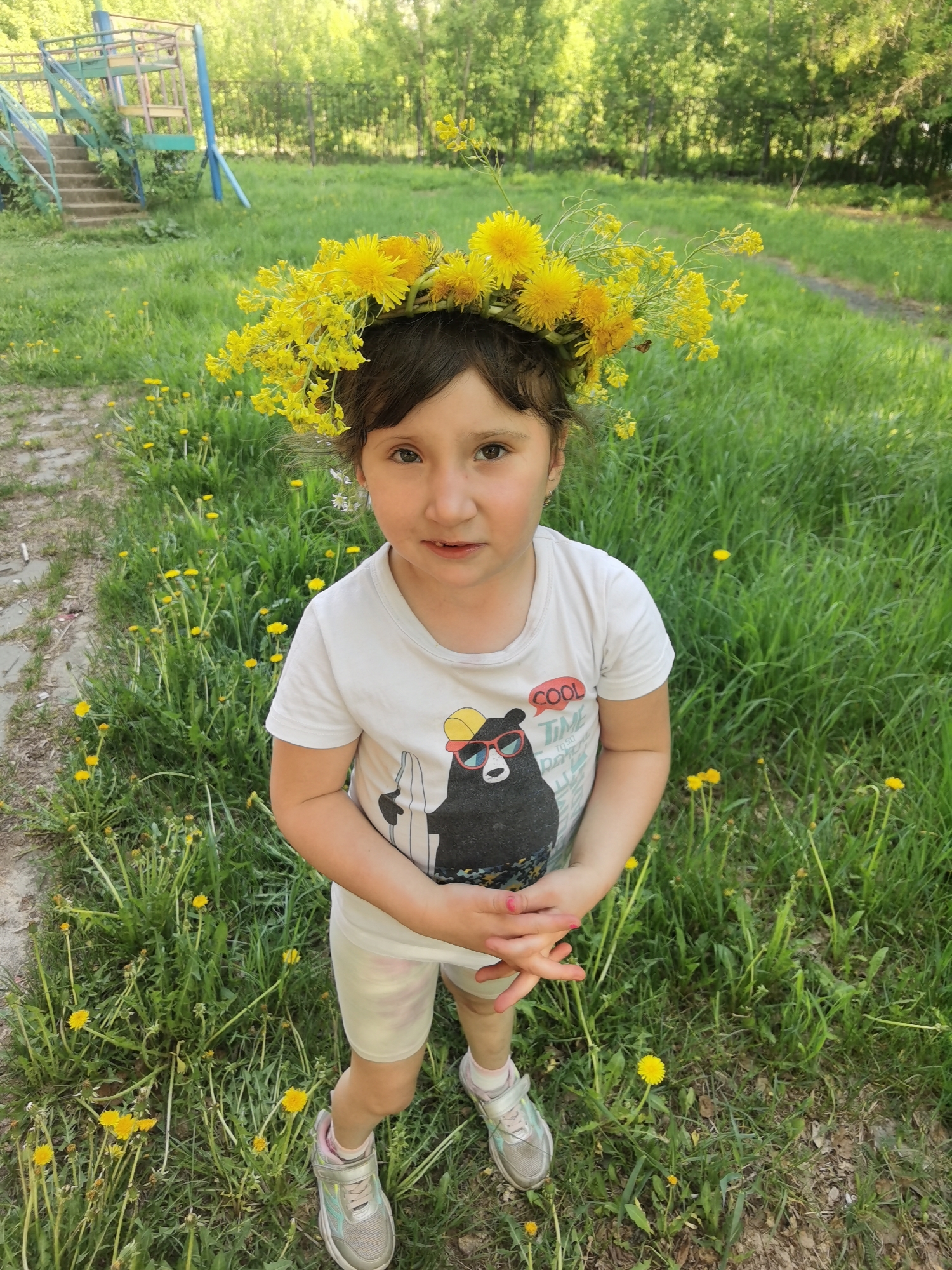 A young girl stands in a grassy field wearing a crown of yellow flowers, likely dandelions. She has dark hair and is wearing a white t-shirt with a cartoon bear on it and light-colored shorts. She stands among numerous dandelions. In the background, trees are visible, along with a simple, elevated play structure. The location is likely Perm, Russia, given that information was specified in the prompt.