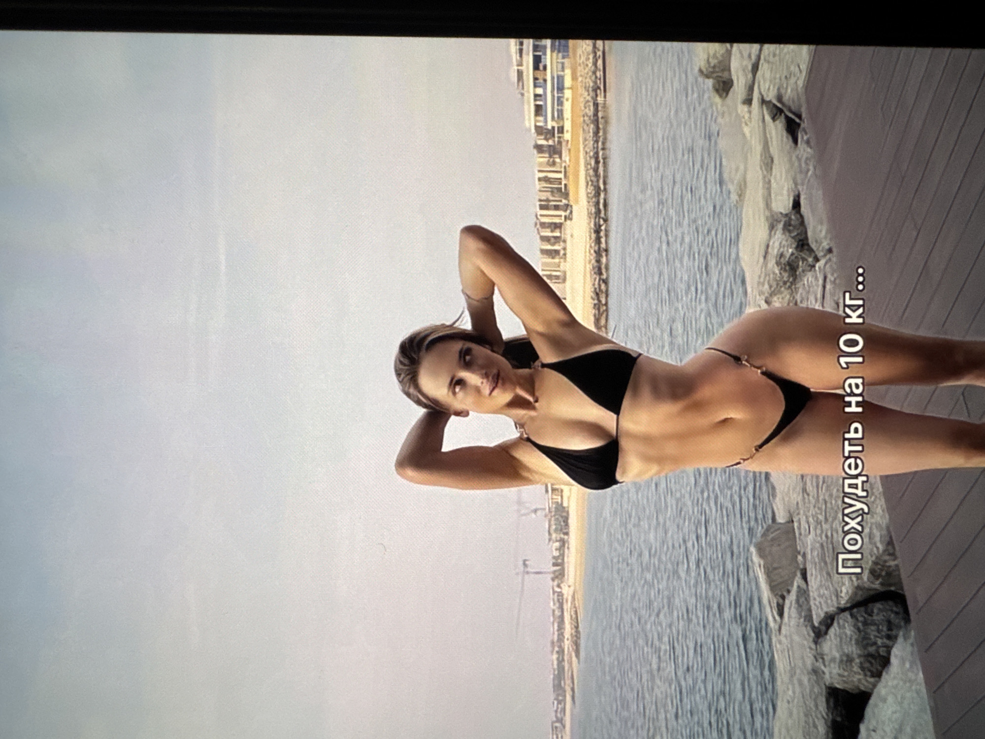 A woman wearing a black bikini is lying on her side near the sea, posing for a photo. The backdrop features buildings in the distance, a gray sky, and a wooden walkway. There is text in a non-English language overlayed on the image. The scene does not appear to be set in Yerevan, Armenia.