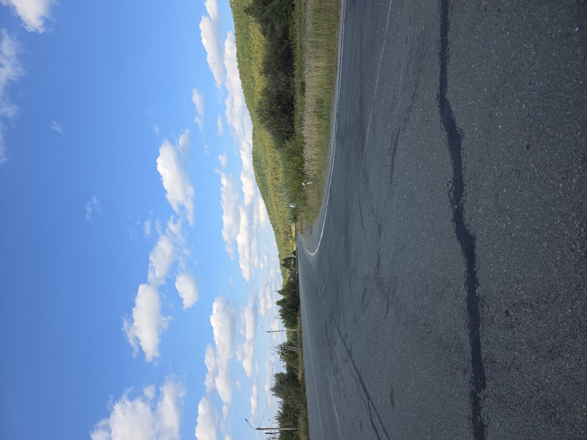 The image depicts an asphalt road curving through a landscape with a dense green forest on a hill to the right, under a bright blue sky with scattered white clouds. The road surface shows some signs of wear, including dark patches that appear to be asphalt repairs or stains. On the left side of the road, there are streetlights and various trees and vegetation. The scene is a scenic view of a road in a natural setting, likely in or near the city of Karabash, Russia, given its geographical context. There are no people or vehicles visible in the image, focusing the attention on the road and the surrounding environment.