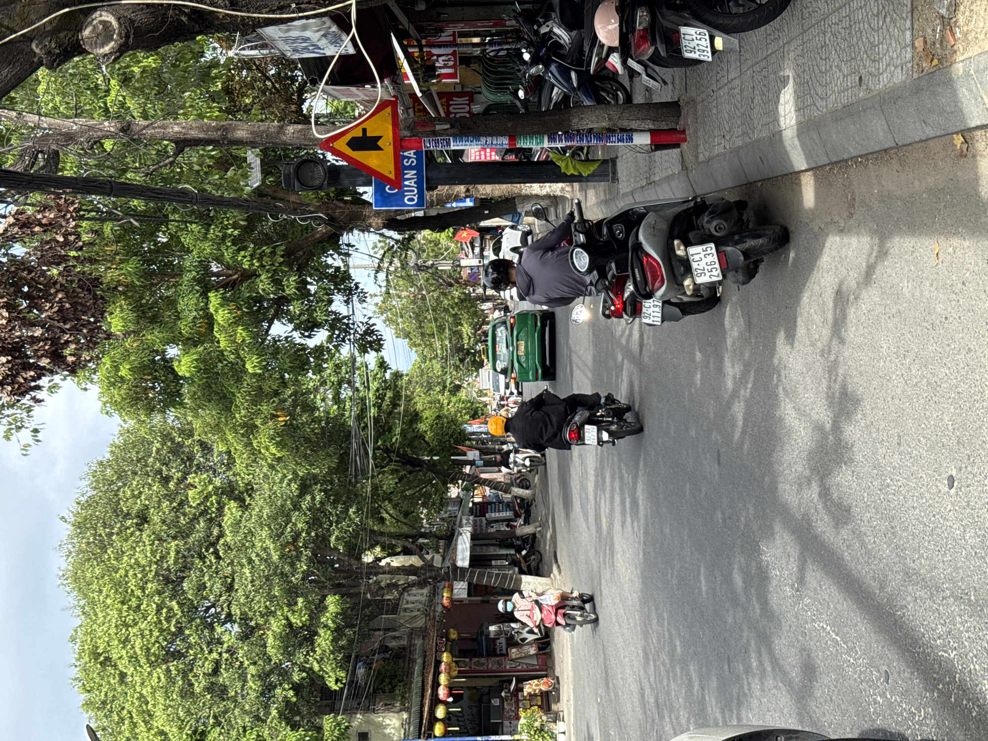 The image depicts a street scene in Hoi An, Vietnam. In the foreground, a gray scooter with license plate "92-C1 256.35" is parked on the right side of the road, facing away from the viewer. To its right, another scooter, also facing away, has a license plate that reads "92-C1 392.56". Further back on the road, a person in a black jacket and a yellow helmet rides a dark-colored motorcycle. Ahead of them, a green taxi is visible in the middle of the road, followed by other vehicles. On the left side of the street, a person wearing a pink shirt and a blue face mask rides a pink scooter. The street is lined with buildings and trees. A traffic light and a yellow triangular warning sign with a black arrow indicating a junction are mounted on a pole. The sky is overcast.