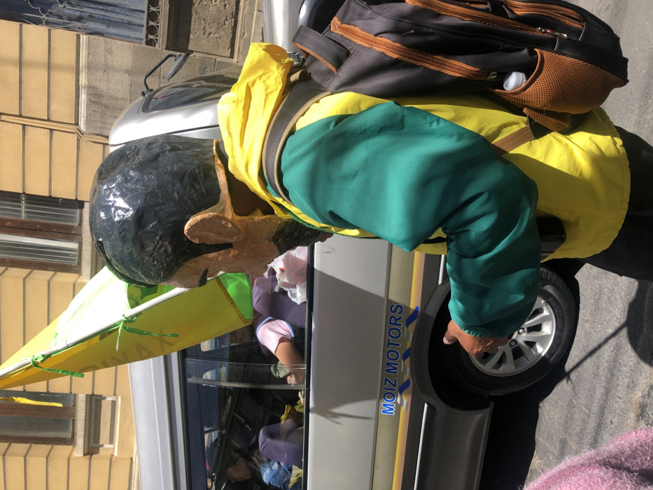 A large papier-mâché head wearing a yellow and green jacket stands next to a van with "Moiz Motors" on its side. The head has dark hair, large ears, and a protruding nose. A yellow flag is attached to the head. A black backpack is worn over the jacket. The interior of the van is visible through the window, revealing a cluttered scene. The scene is outdoors on a sunny day.
