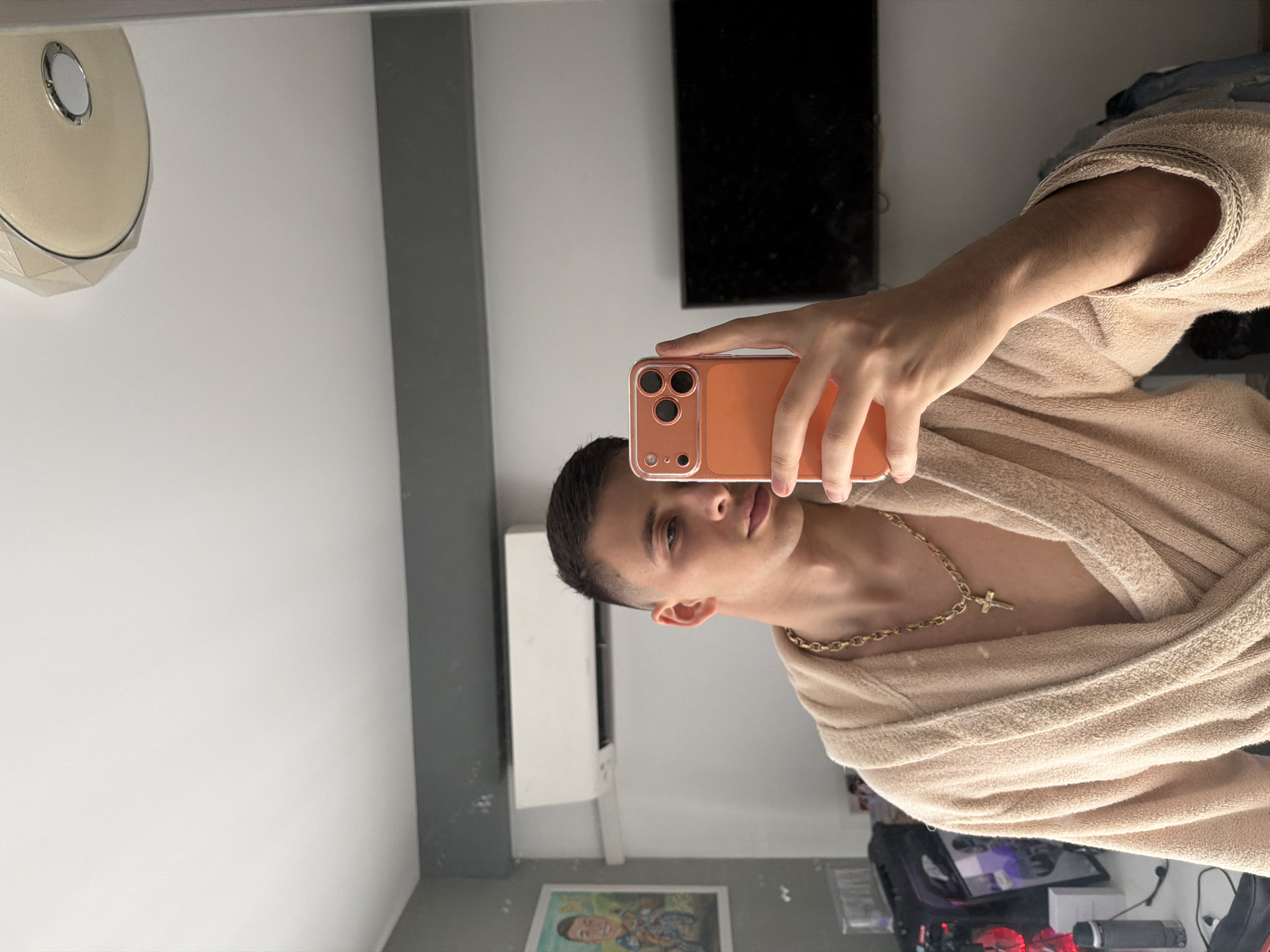 A person is taking a selfie in front of a mirror. The person is wearing a tan robe and a gold chain with a cross pendant. The person is holding an orange phone. The background includes a black rectangle (possibly a TV screen), a light fixture, an air conditioning unit, and a framed picture. The location in Plovdiv, Bulgaria is not discernible from the image.