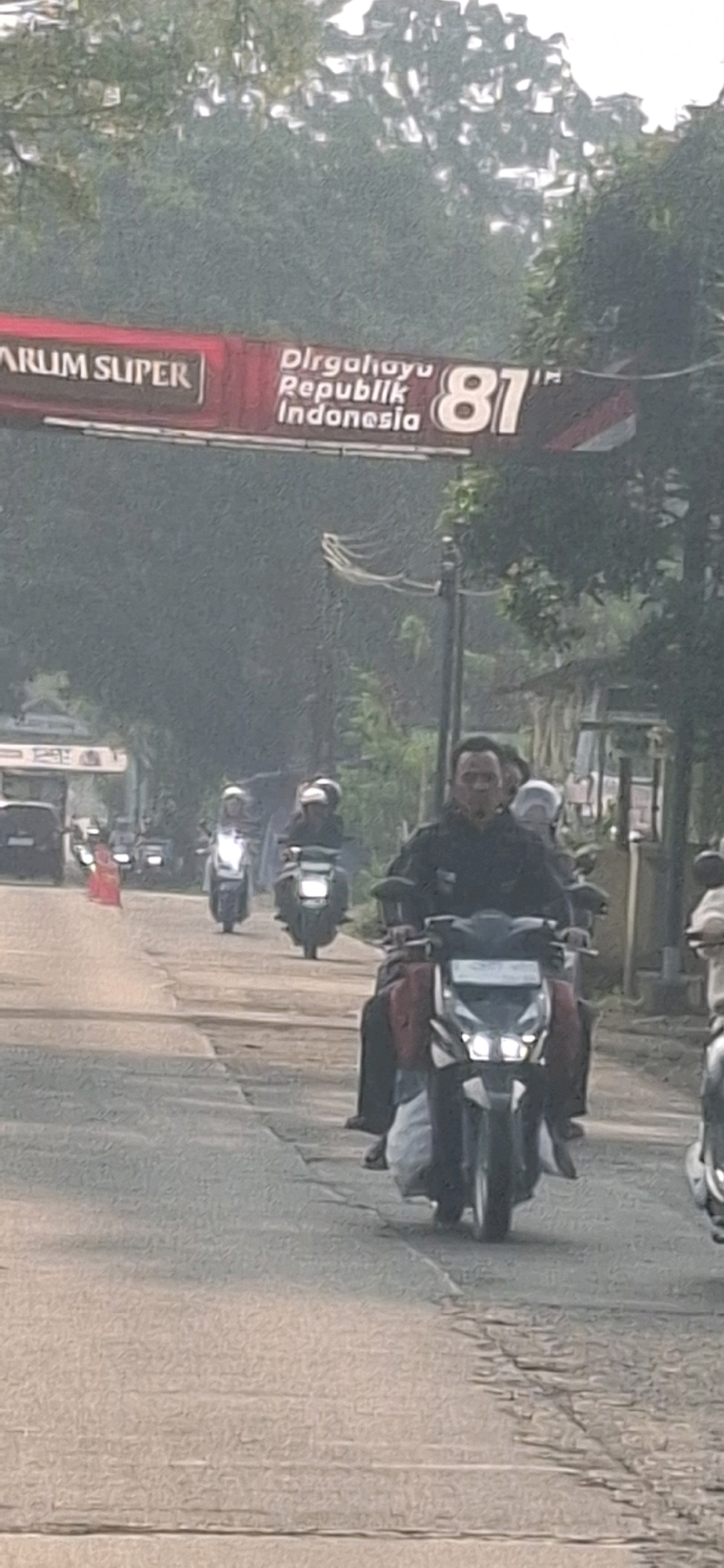 In Sukasari, Indonesia, a street scene unfolds with several motorcycles on a paved road, suggesting urban or semi-urban traffic. Overhead, a banner reads "PARUM SUPER" and "Dirgahayu Republik Indonesia 81", indicating a celebration for Indonesia's 81st Independence Day. The road shows signs of wear, with visible cracks. On the right side of the image, a man in a black jacket is riding a motorcycle, and a red-clad figure is visible further down the road. Other individuals on motorcycles are also present, some wearing helmets. The environment appears somewhat hazy, possibly due to fog or pollution.