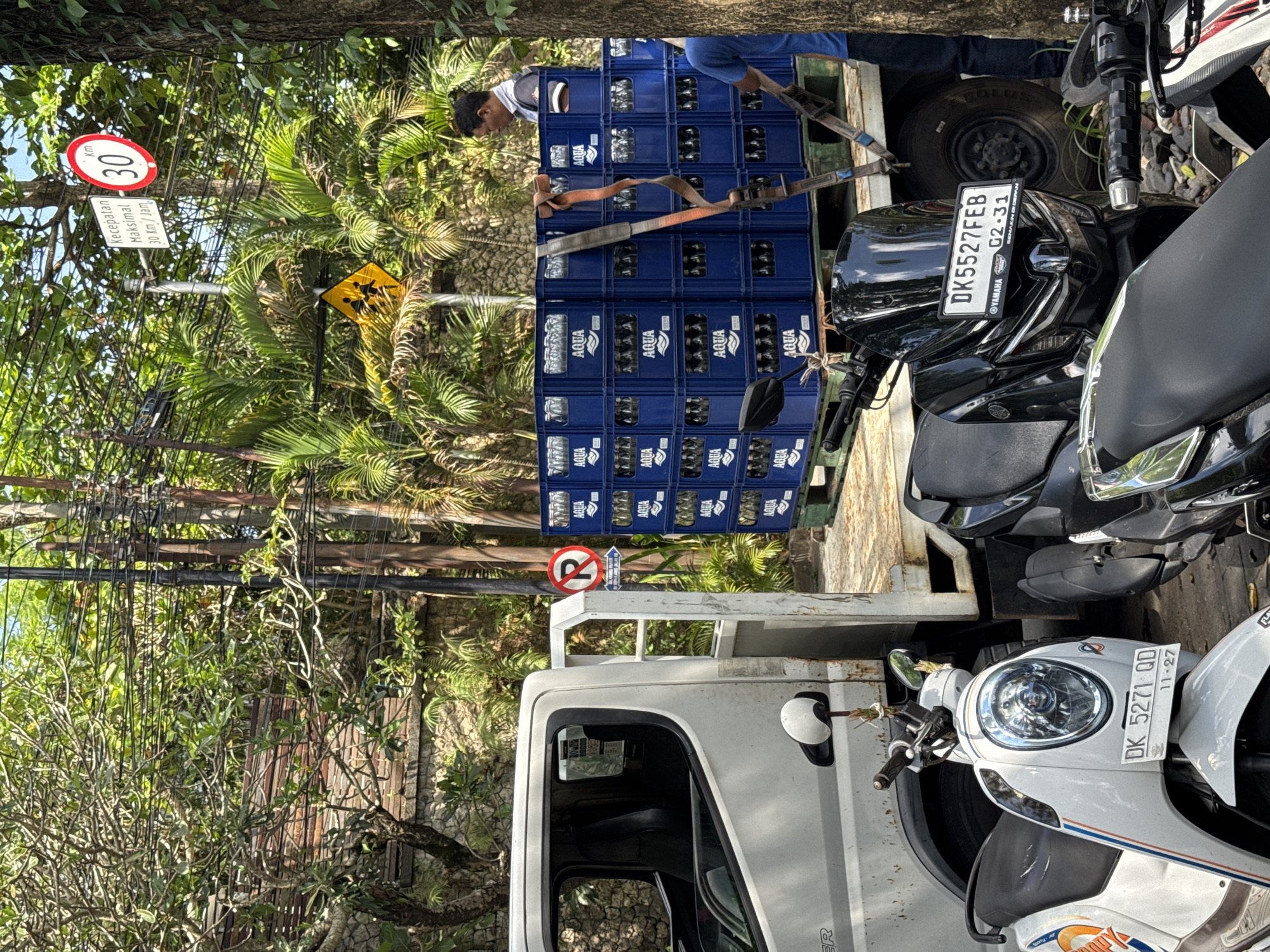 Here is a detailed description of the image:
**Overview:** The image captures a street scene in Kuta Selatan, Indonesia. The scene is dominated by a white delivery truck loaded with stacks of blue Aqua water bottle crates. A person, partially visible, is handling the crates. Two parked motorbikes obscure the truck. Traffic signs are present in the background.
**Specific Details:**
* **People:** A single person is visible working on the truck, likely involved in loading or unloading the water crates.
* **Objects:** The prominent object is the delivery truck filled with Aqua water crates. Two motorbikes are parked next to the truck. Several traffic signs are visible, including a speed limit sign (30 km/h) and a no U-turn.
* **Actions:** The person appears to be involved in handling the Aqua crates on the truck.
* **Environment:** The environment features tropical foliage, suggesting the location is in a warm, humid climate. The presence of traffic signs and vehicles indicates a roadside setting within Kuta Selatan.
* **License plates:** The black motorcycle has a license plate that reads "DK5527FEB 02-31". The white motorcycle shows "DK 5271 QD 11-27".