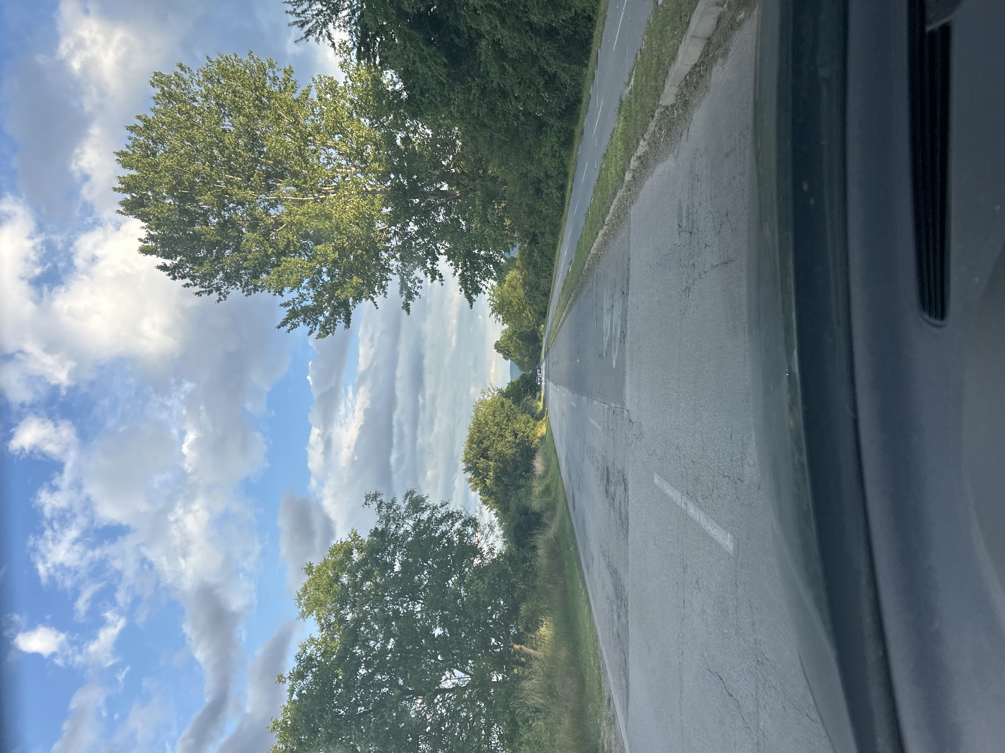 An outdoor, bright daylight scene depicts a road stretching into the distance, flanked by trees and greenery under a partially cloudy blue sky. The image appears to be captured from inside a vehicle, with a dark, possibly grey or black, car pillar or frame visible along the right side.
The asphalt road, showing some signs of wear and cracking, has a white dashed line marking its edge or lane. Lush green trees with full foliage line both sides of the road, extending into the background. Some areas of dry grass are also visible along the road's verge. The sky is a mix of clear blue and scattered white cumulus clouds, with some darker, more elongated cloud formations appearing in the distance towards the left. In the far distance along the road, faint shapes of what might be other vehicles can be discerned.
The setting suggests a rural or semi-rural road. The time of day is clearly bright daylight, indicating good weather conditions. No specific individuals or explicit activities are shown, other than the implied movement of the vehicle from which the photograph was taken. No visible text is present. The scene captures a moment of travel through a verdant landscape, consistent with an area like Kazanlak, Bulgaria.