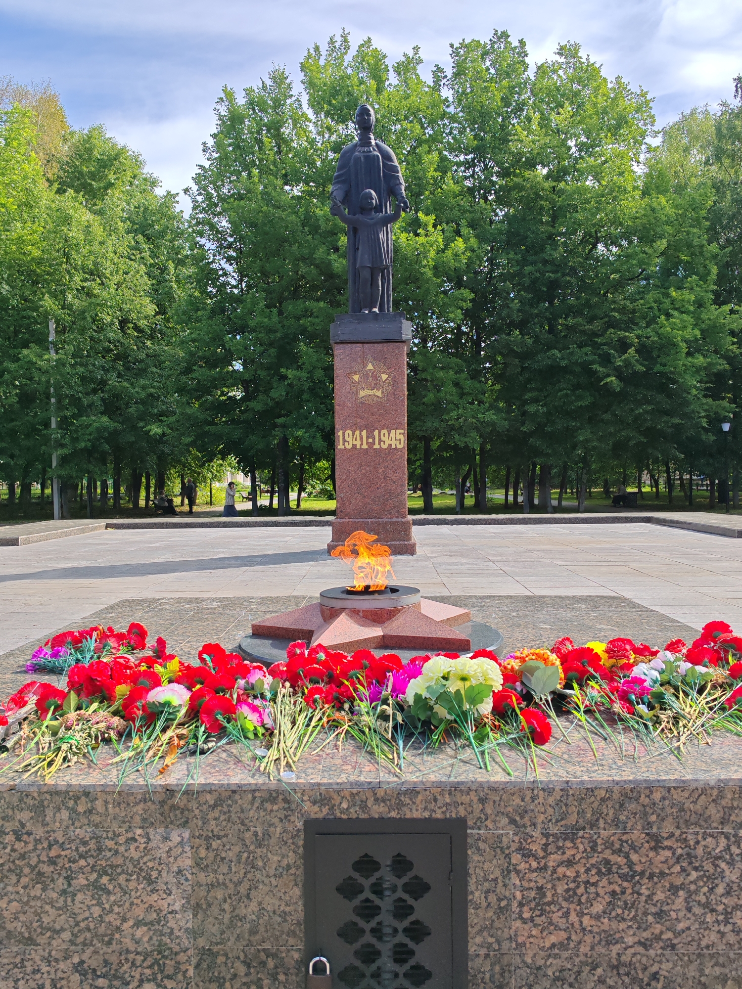 The image depicts a memorial monument in Buy, Russia, featuring a bronze statue of a woman holding a child's hand, standing atop a red granite pedestal inscribed with "1941-1945." A perpetual flame burns in front of the monument, and an assortment of colorful artificial flowers, predominantly red, are laid out at its base. The monument is situated in a paved public square, surrounded by a lush green park with numerous trees. In the background, a few individuals are visible, appearing to be visitors or residents enjoying the park. The sky is partly cloudy, suggesting it is daytime. The overall scene conveys a somber and respectful atmosphere, commemorating a significant historical period.