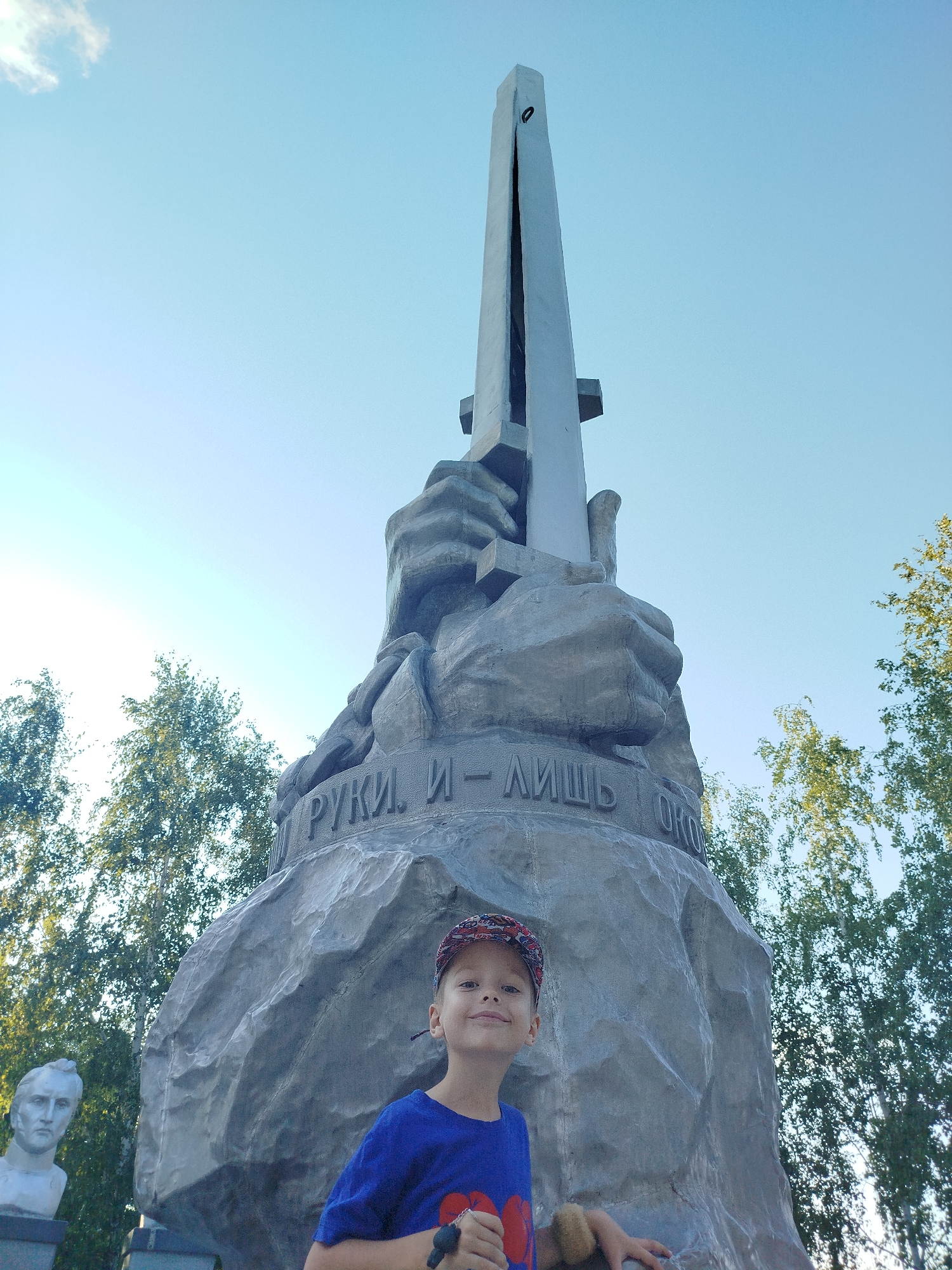 A low-angle shot captures a young boy with light skin, brown hair, and blue eyes standing in front of a large, grey stone monument under a clear blue sky with a few wispy clouds. The boy is wearing a blue t-shirt with a red graphic and a patterned baseball cap. His left arm is visible, resting on the monument, and he is holding a small, light brown stuffed toy. To the left of the boy, a white bust of a man is visible on a pedestal. The monument itself is abstract, featuring large, stylized stone hands holding up a tall, metallic obelisk that is split down the middle. Engraved text is visible on the monument's surface. Surrounding the monument are green trees, their foliage visible against the bright sky. The scene appears to be outdoors in a park or public space. The monument is the "Monument to the Defenders of the Fatherland" located in the city of Yalutorovsk, Russia.