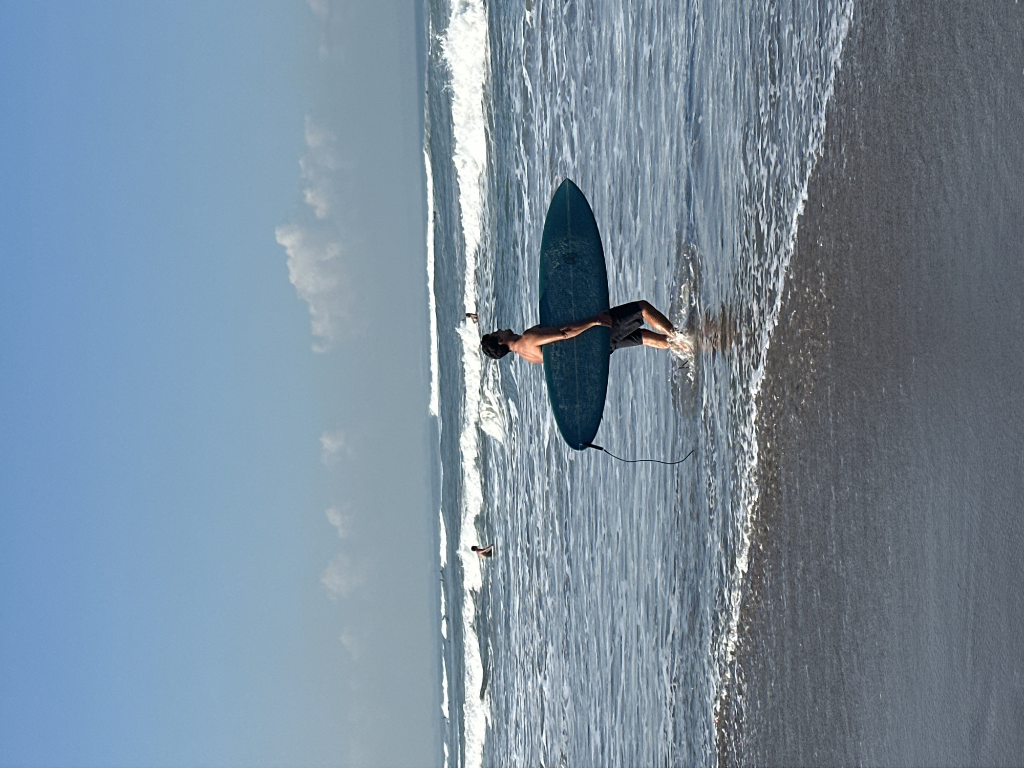In North Kuta, Indonesia, on a beach with a clear blue sky and scattered white clouds, a man is walking in the ocean, carrying a dark teal surfboard. He is wearing black swim trunks and is submerged up to his knees in the water. The surfboard is positioned vertically, with the tail end resting on the ocean floor and the nose pointed upward. A black leash extends from the surfboard's tail. Further out in the ocean, other individuals are visible, some appearing to be in the water and others possibly on surfboards. Waves are breaking in the background, creating white foam on the surface of the water. The foreground shows the wet sand of the beach with gentle waves lapping at the shore.