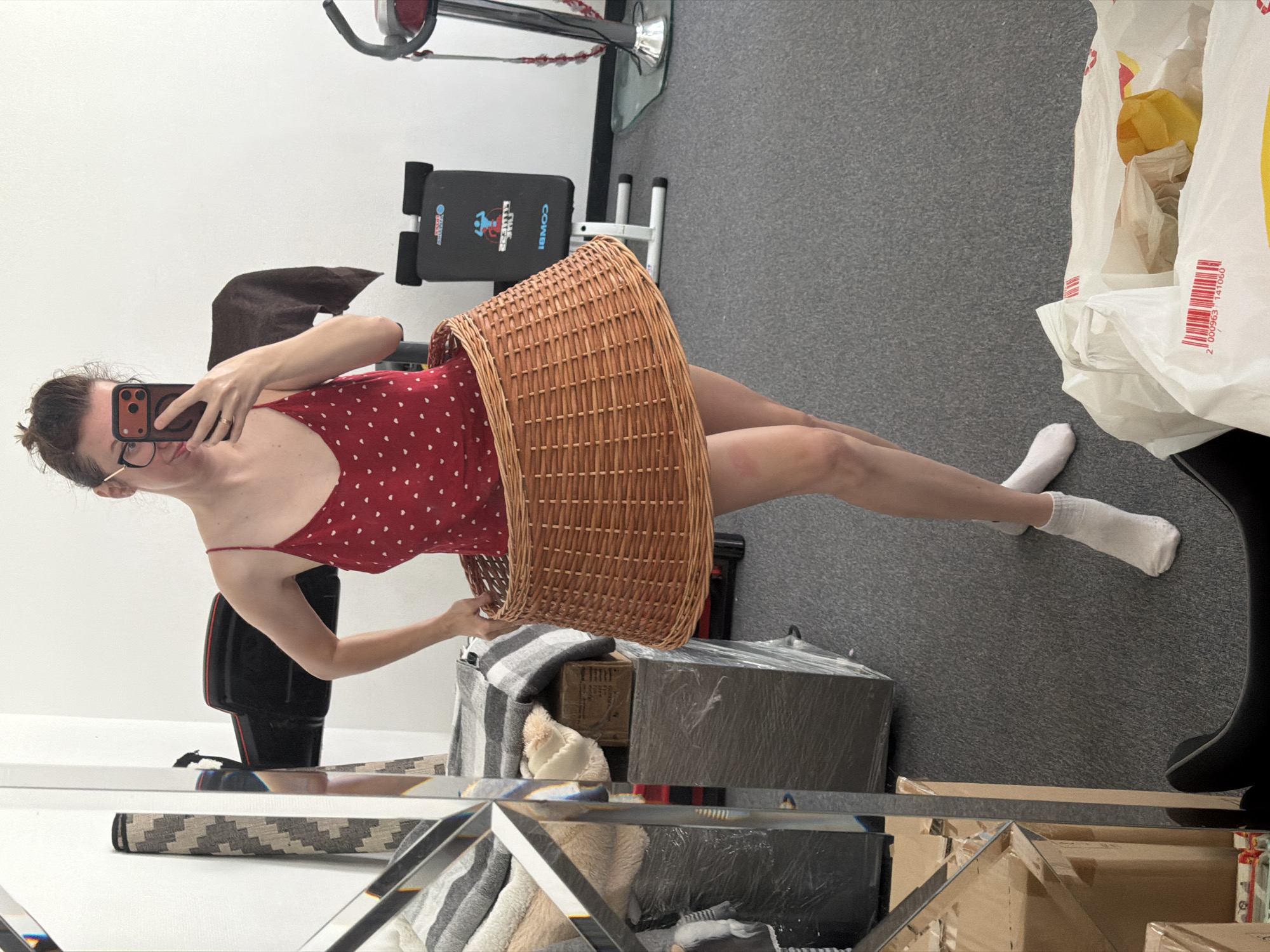 A woman stands in what appears to be a home gym or storage area, holding a smartphone and taking a mirror selfie. She is wearing a red dress with white polka dots and has a large wicker basket around her waist, resembling a skirt. The environment includes gym equipment, storage boxes wrapped in plastic, and a mirror reflecting part of the scene. The location is not specified as being in Bila Tserkva, Ukraine.