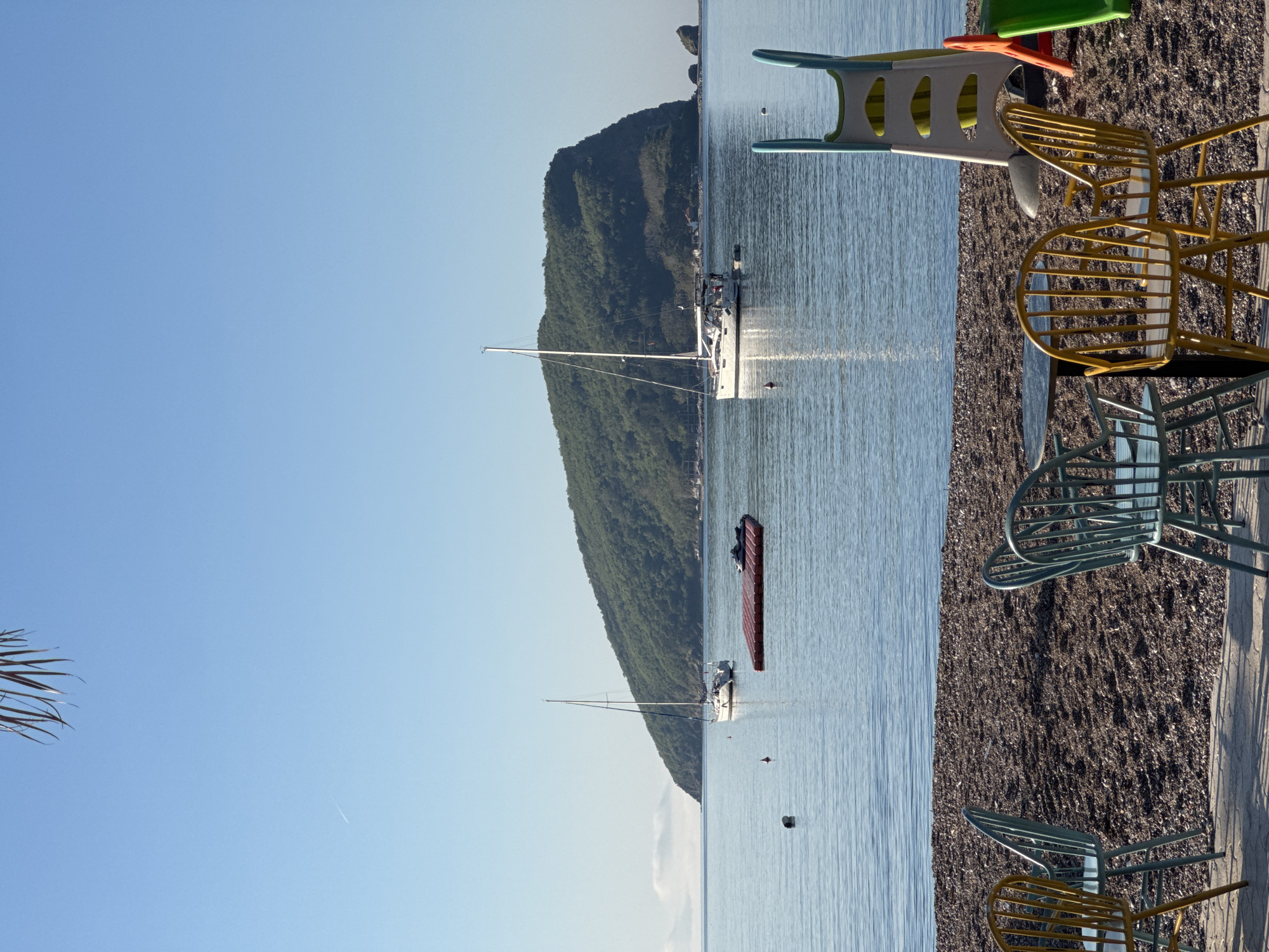 The image shows a landscape with a body of water, likely the Adriatic Sea, adjacent to a landmass covered in dense greenery. The location is suggested to be near Budva, Montenegro, a coastal city known for its beaches and mountainous backdrop.
Several sailboats are anchored in the water. One has its mast clearly visible. A red floating object is also visible. The water is relatively calm, reflecting the sunlight. The sky is clear blue.
The foreground consists of a pebble beach and outdoor furniture, specifically colorful chairs.