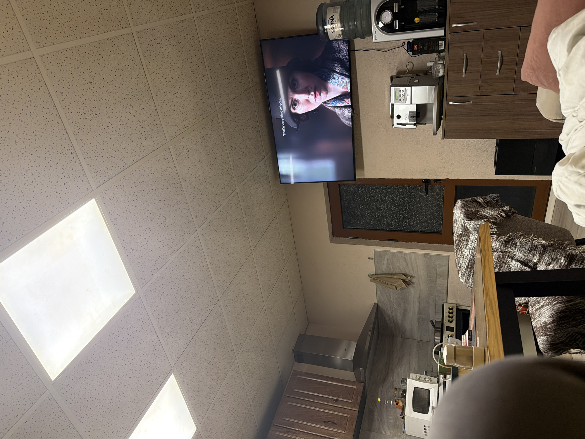 The scene depicts the interior of a room in Panagyurishte, Bulgaria. A television is mounted on the wall displaying a person's face with the subtitles "That's very kind of you." Adjacent to the television is a water dispenser and a coffee machine on top of a set of brown cabinets. The room has a drop ceiling with square lights. A doorway leads to a kitchen area, visible with gray tiles, a range hood, and a microwave. A table and chair are in the foreground.