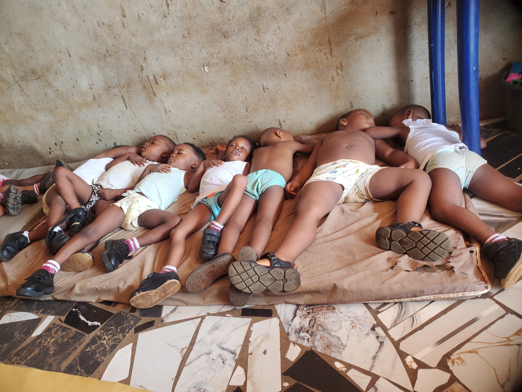 Six children are lying side-by-side on a thin, light brown mattress on a marble tile floor. The children are arranged from left to right, their bodies parallel to each other. All of them are wearing shoes and socks. Most of the children are wearing light-colored shirts and diapers or shorts. Their eyes are closed, and they appear to be sleeping. The background is a light-colored wall with paint splatters and other imperfections. Two blue metal posts are visible behind the children on the right. The photograph was taken in Onitsha, Nigeria.