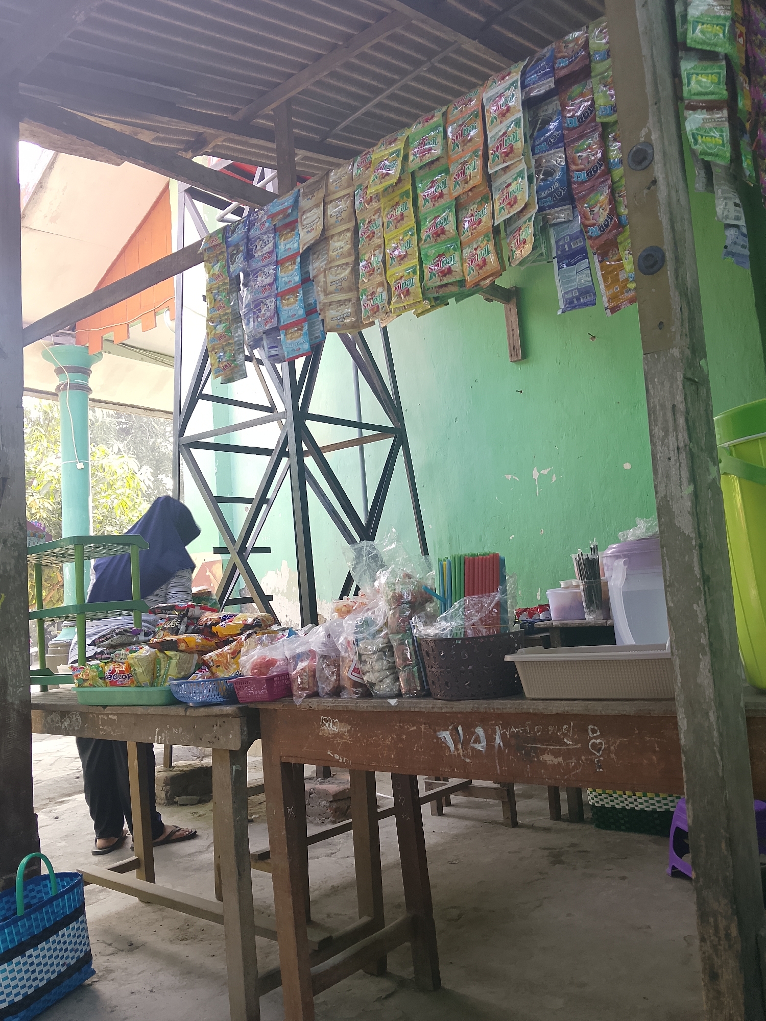 This is an outdoor market stall in Kepuh, Indonesia. A vendor, partially obscured by a blue headscarf and dark clothing, stands behind a wooden table laden with various packaged goods, including snacks and small sachets of products. Behind the table, a green wall serves as a backdrop, and above, a corrugated metal roof is supported by a rustic wooden structure. Packets of merchandise are hung from the roof, displaying vibrant colors and branding. The ground is paved with concrete, and a blue and white woven basket sits on the left side of the frame. The scene suggests a small, local business operating in an open-air environment.