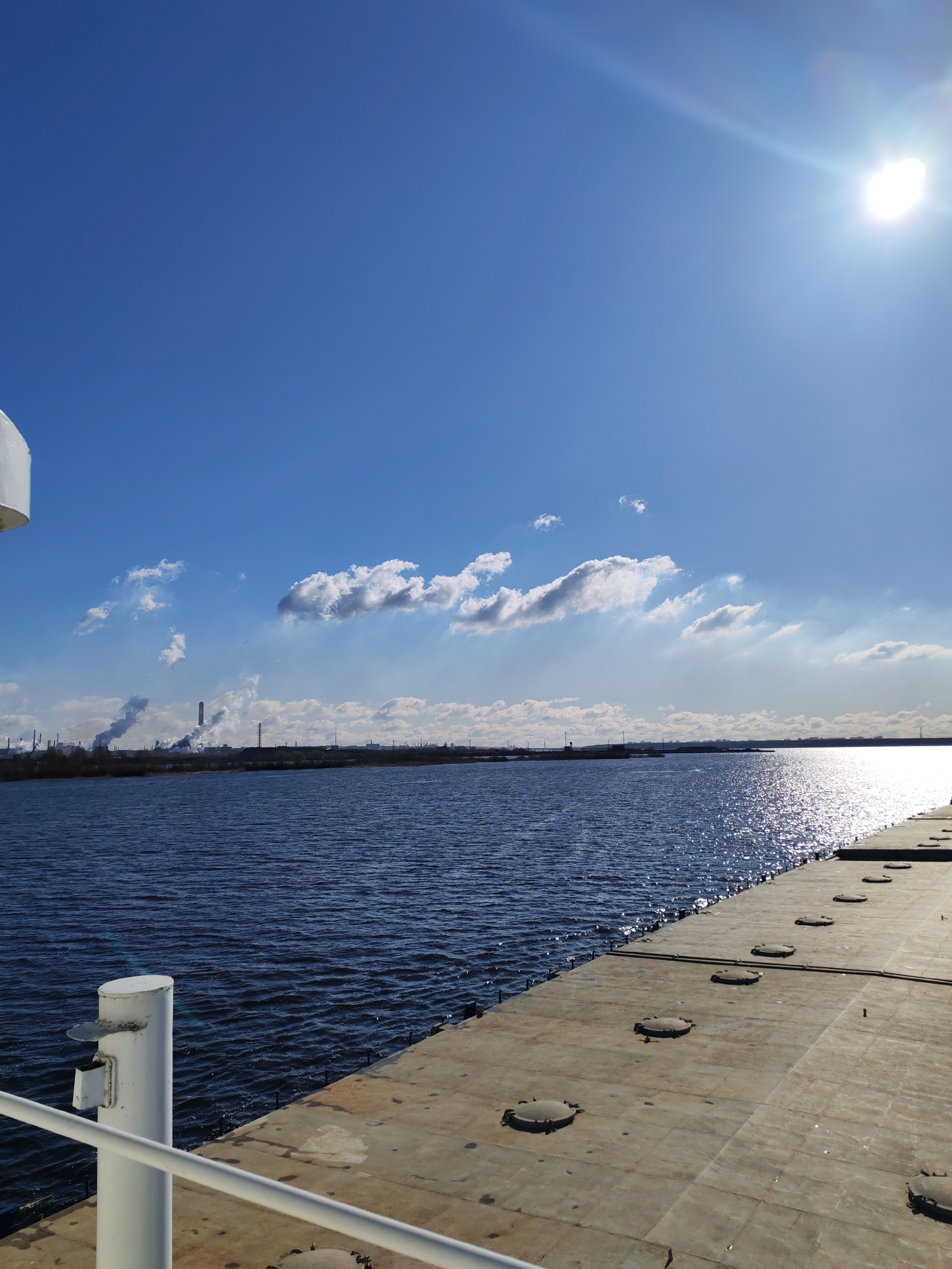 The image shows a view of the river and sky from what appears to be a deck. The sky is blue with some white clouds and the sun is bright. On the horizon, there is an industrial area with visible smoke stacks. The river is dark blue with sunlight reflecting off its surface. The deck is made of metal plates. The image may be taken from a ship or a dock in Суда, Russia, but this cannot be verified.