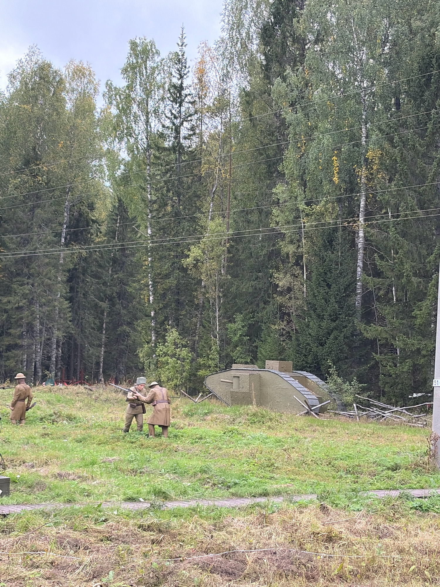 In Kukhtoozerskiy, Russia, a historical reenactment takes place in a grassy field bordering a dense forest. Three individuals dressed in WWI-era military uniforms are present. One person stands to the left, facing right. Two others are in the center, engaged in an interaction where one appears to be handing a rifle to the other, who is holding it. A model of a WWI tank, painted olive green, is positioned to the right of the central figures. The environment consists of tall trees, green grass, and some dry, dead grass. Overhead power lines are visible. The sky is overcast.