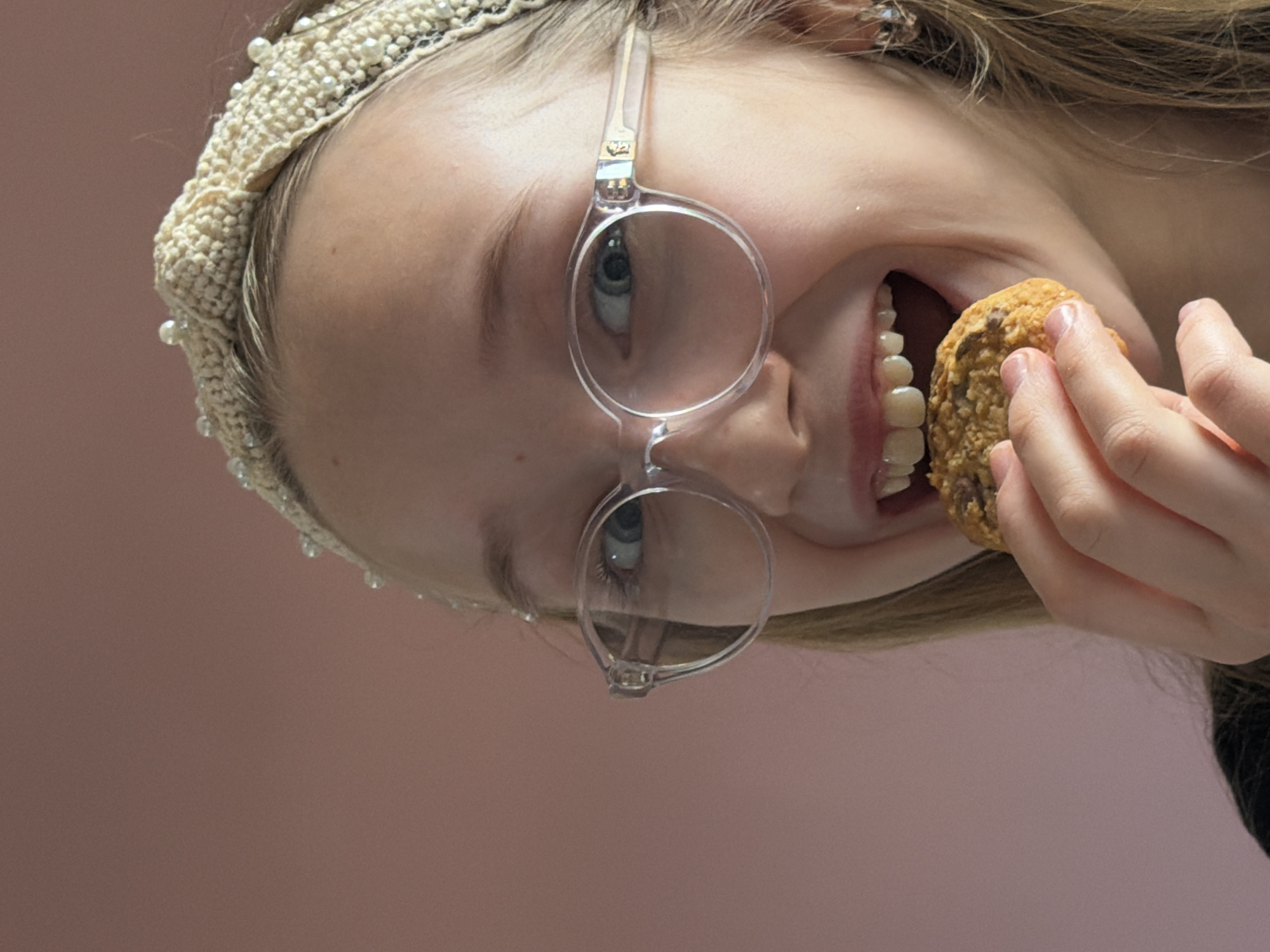 A close-up portrait taken in Dreiliņi, Latvia, depicts a young individual with fair skin, light-colored hair, and round, clear-framed eyeglasses. The subject wears a cream-colored, pearl-beaded headband. The individual's mouth is open, revealing upper and lower teeth, as a light brown cookie with visible inclusions (such as chocolate chips) is held to the mouth by the right hand, positioned for a bite. The eyes are light blue and gaze directly forward. A small, metallic earring is visible in the right earlobe. The background is a solid, muted pink wall, indicating an indoor setting.