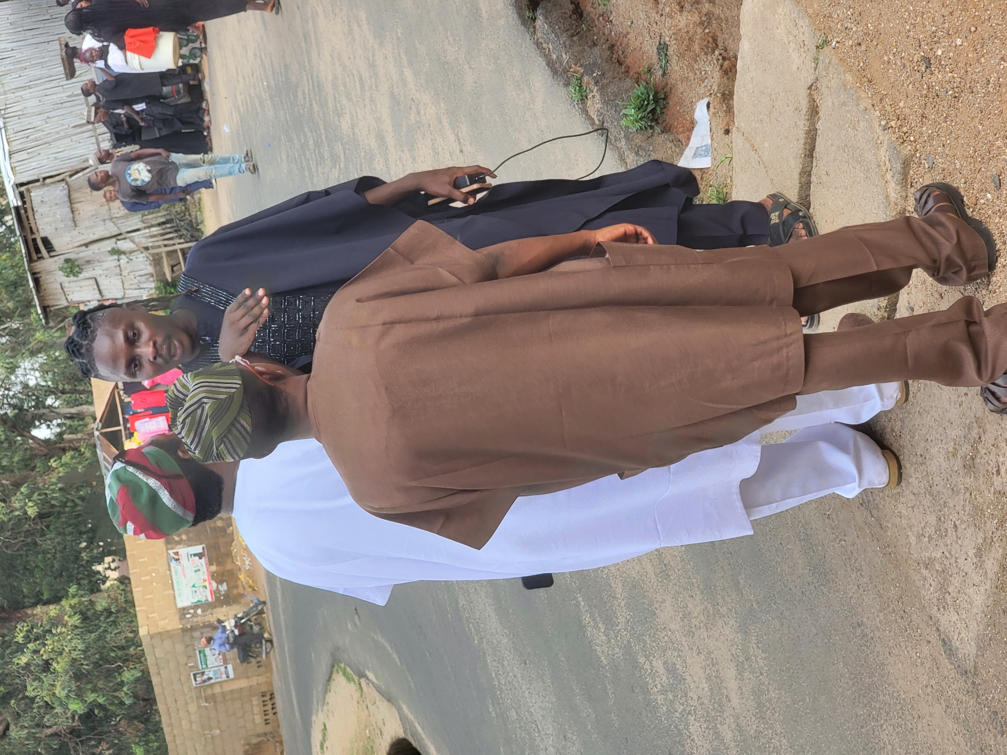 The image shows two men standing outside in Jos, Nigeria. One man is wearing a white robe with a brown outer garment and a green-striped hat. The other man is wearing a dark blue robe. Both men are wearing sandals or shoes. In the background, there are buildings and other people. The ground is paved. The presence of nearby foliage suggests an urban environment, likely on a road or sidewalk.