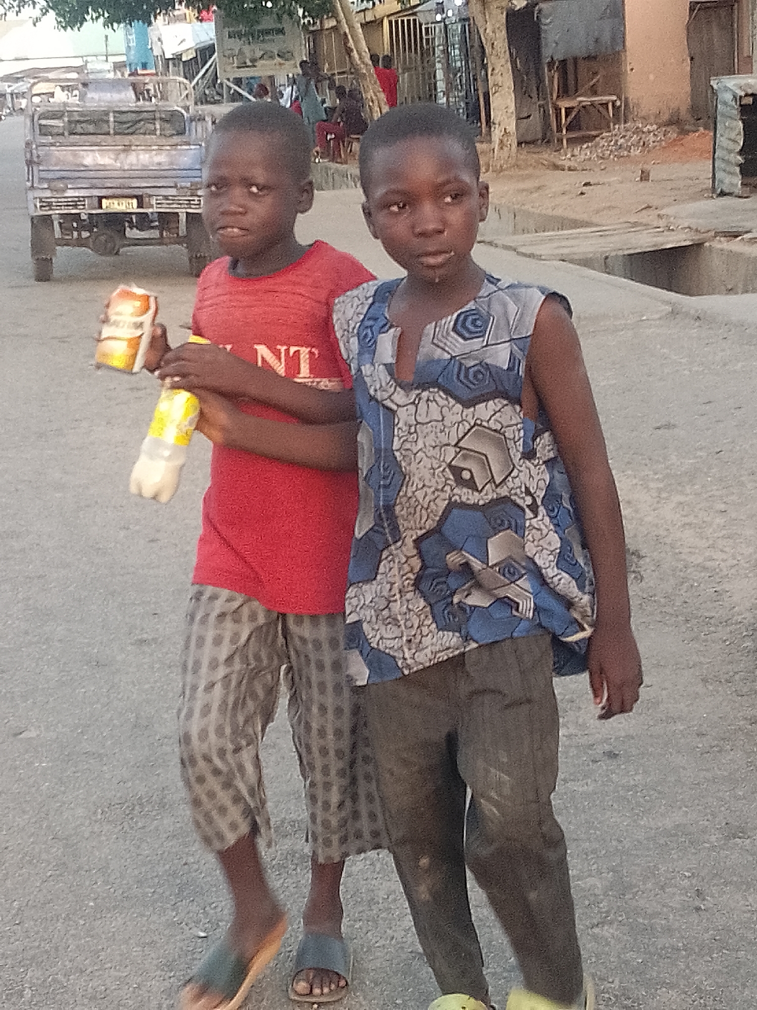 Two young Black boys are standing on a paved road in what appears to be an outdoor market or residential area in Pantame, Nigeria. The boy on the left is wearing a red t-shirt and patterned trousers, and is holding a can of drink and a bottle of liquid. The boy on the right is wearing a sleeveless blue and white patterned top and dark trousers, and has his arm around the other boy's shoulder. Both boys appear to be looking towards the viewer. In the background, there is a truck, various buildings, and other people. The lighting suggests it is either early morning or late afternoon.
