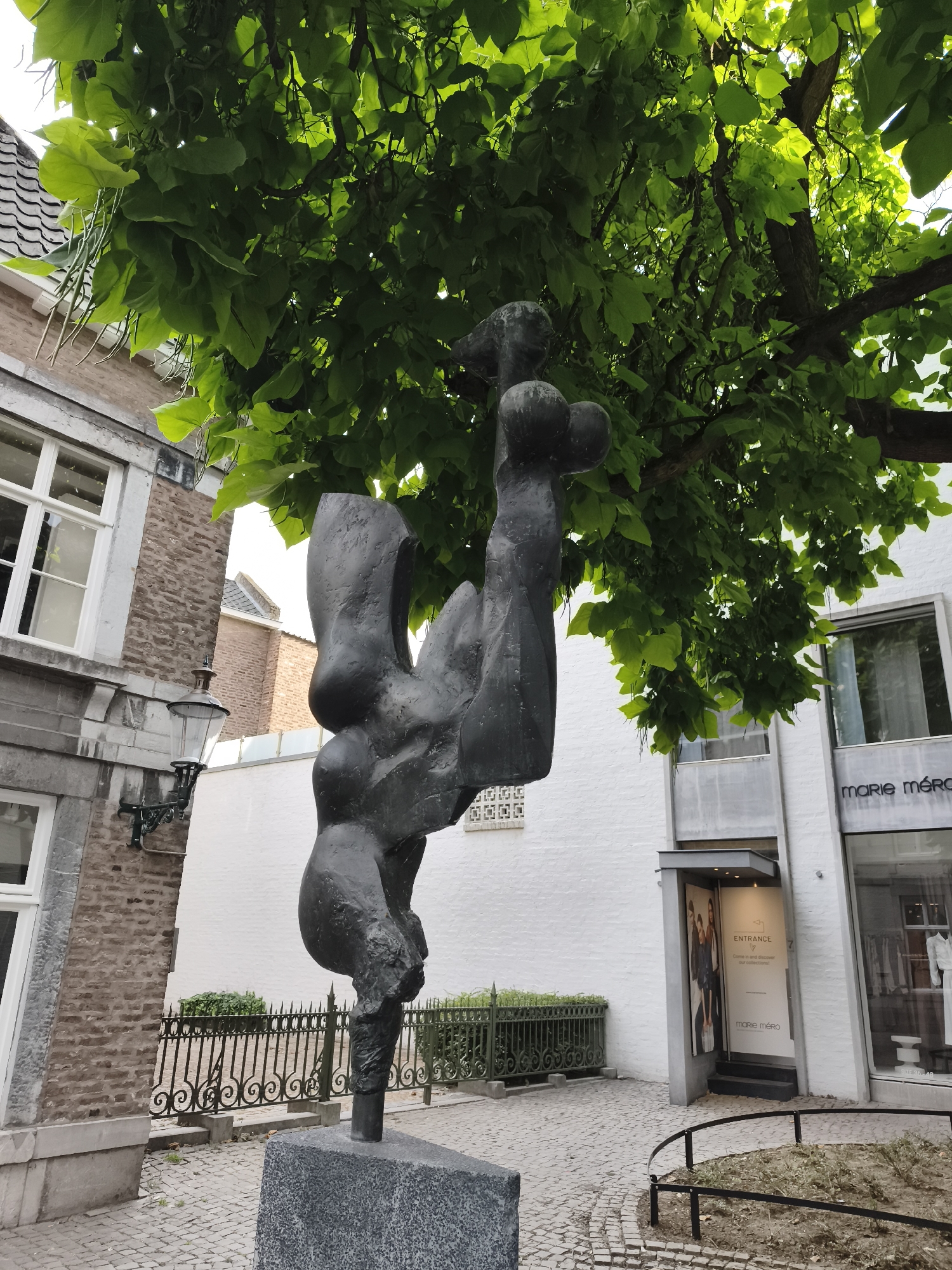 This is a medium shot of a black, abstract metal sculpture located on a cobbled street in Maastricht, Netherlands, under a tree with bright green leaves. The sculpture is supported by a dark gray stone base. To the left of the sculpture is a building with a stone facade and white window frames, and a vintage-style lamp attached to the wall. To the right of the sculpture, there is a white building with a glass shopfront displaying clothing, and a sign that reads "MARIE MÉRO". The entrance to the shop is visible with a banner that says "ENTRANCE". A black wrought-iron fence with a decorative pattern encloses a small patch of greenery behind the sculpture. The sky is visible in the upper right corner, appearing overcast.