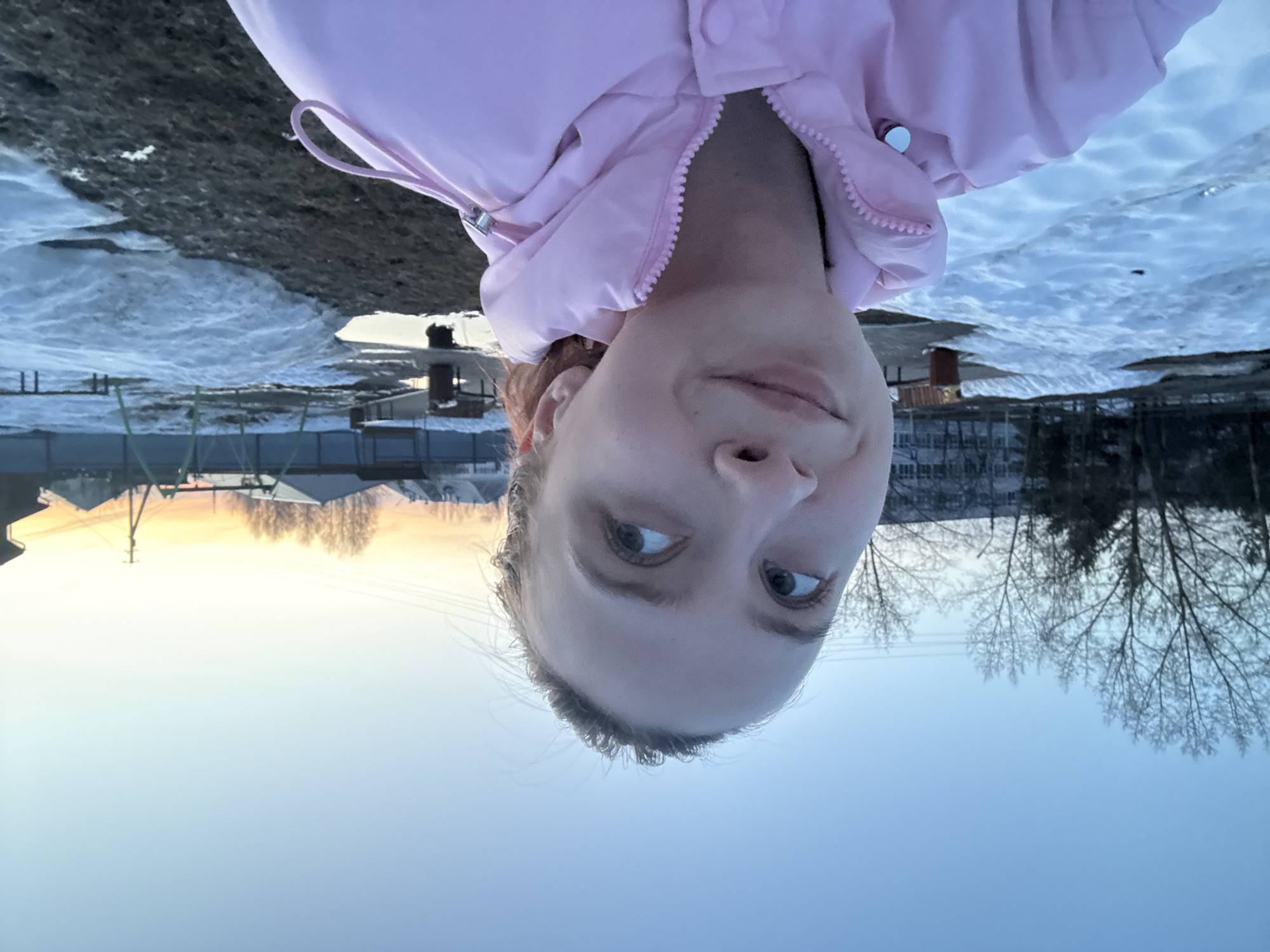 A selfie shows a fair-skinned person with light brown hair wearing a pink puffer jacket. The background depicts a winter scene with remnants of snow on the ground. Houses and leafless trees are visible against a sky that transitions from light blue near the bottom to a pale orange at the horizon. The image was taken in Прикамский, Russia, as evidenced by the architecture and environment.