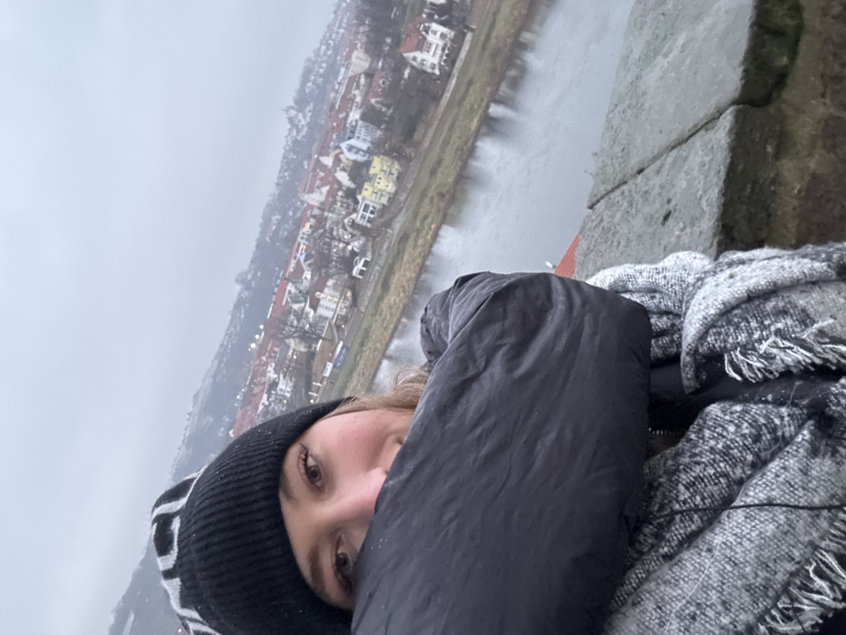 An image, captured in portrait orientation and rotated 90 degrees counter-clockwise, features a young woman in the foreground against a scenic backdrop of a European town and river.
The woman is positioned on the left side of the frame (when viewed correctly), with her face partially obscured by her dark puffer jacket, revealing one eye and a portion of her nose and cheek. She is wearing a black knitted beanie with a white pattern and a textured grey and white scarf with fringe, suggesting cold weather. Her visible eye looks directly at the viewer, indicating a selfie or a close-up photograph.
The background showcases a river flowing through a valley, bordered by a town with numerous buildings featuring diverse roof colors (red, white, yellow) and light-toned facades. Beyond the town, the landscape is hazy and atmospheric, hinting at distant hills or urban areas under an overcast, grey sky. A robust stone wall or parapet is visible on the right side of the image, suggesting the vantage point is from a raised structure overlooking the river and town, likely in Meissen, Germany. The overall atmosphere is cool and cloudy, consistent with the heavy clothing worn by the subject. No discernible text is visible in the image.