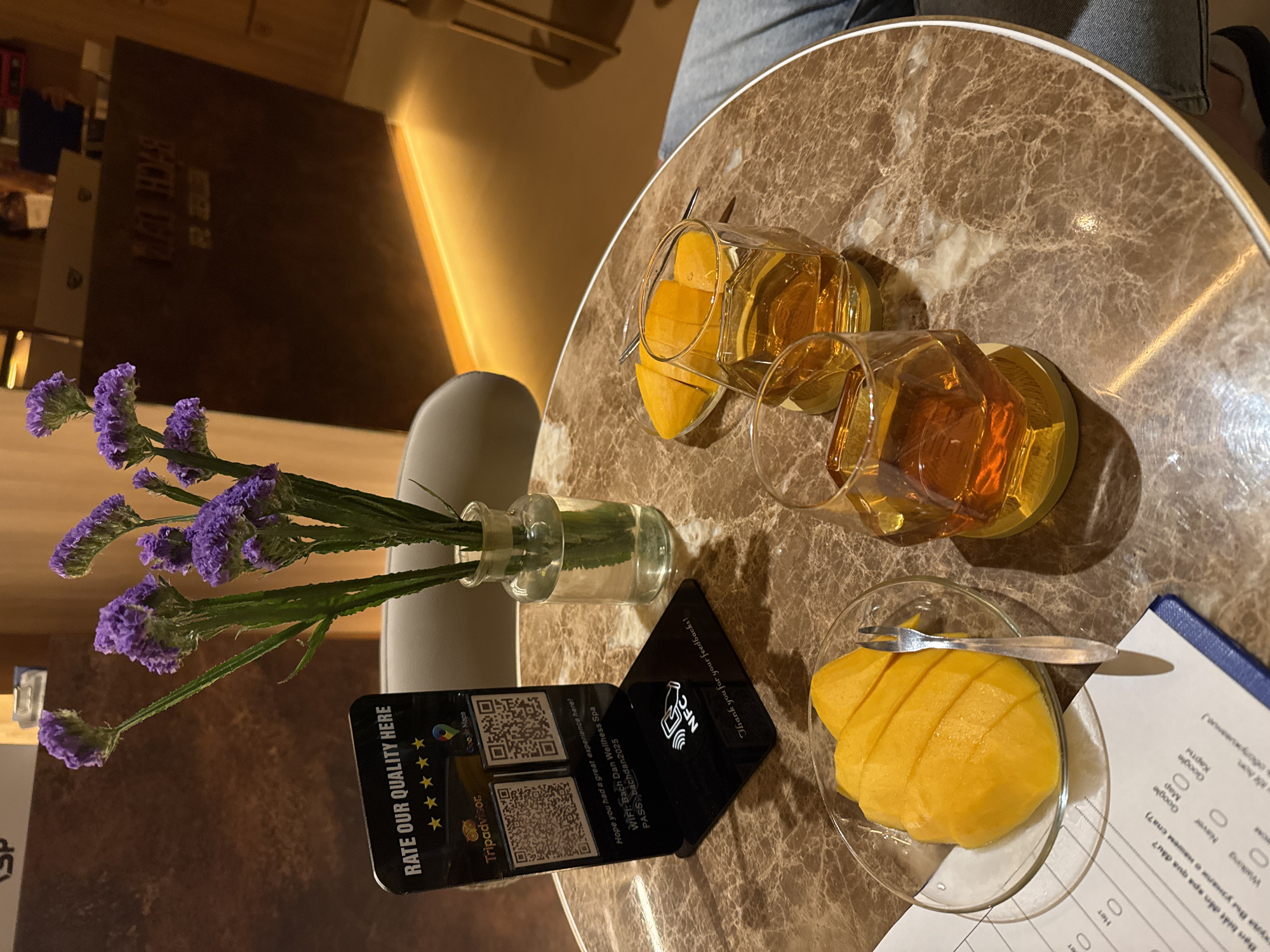Here is a concise description of the image:
The image shows a close-up shot of a round table in what appears to be a cafe or spa. On the table, there are two glasses of what looks like iced tea, a small plate of sliced mango with a fork, a small vase with purple flowers, a black card displaying QR codes and the business's WiFi information (Bach Dan Wellhess Spa, pass: bachdan2025) with Tripadvisor and Google Maps review requests, and a customer service questionnaire. The table has a marble pattern and the setting appears to be in an indoor, well-lit area. An unidentified person is present, partially visible in the top right corner of the photo.