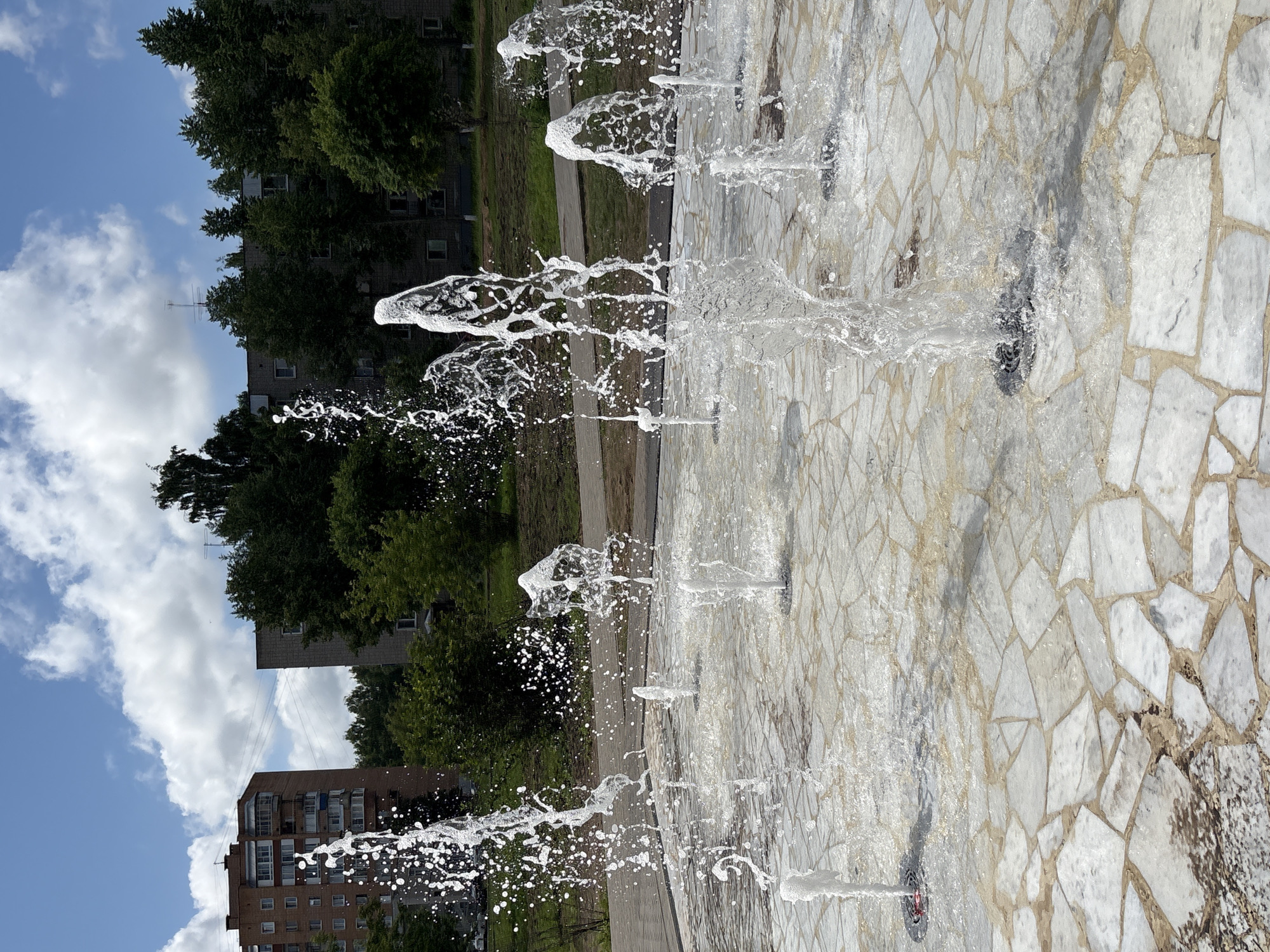 This image depicts a public fountain located in the city of Kirovo-Chepetsk, Russia. The fountain is situated on a paved area with irregular white and beige stone tiles. Several water jets are actively spraying water upwards, creating dynamic, sculptural forms and splashing onto the surrounding tiles. In the background, there are apartment buildings constructed from brick and grey concrete, along with lush green trees and a blue sky with white clouds. The scene appears to be a typical urban outdoor space on a clear day.