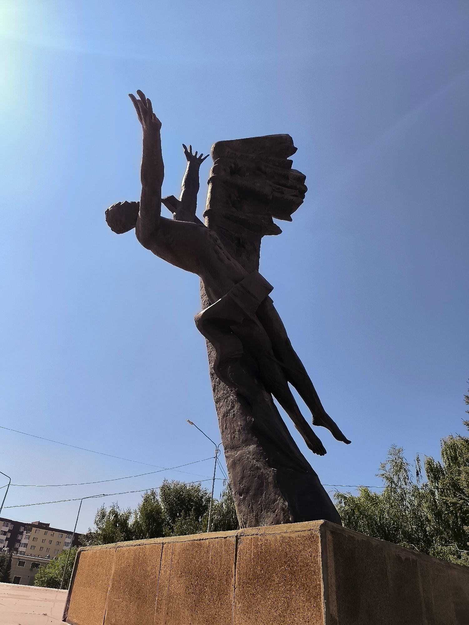 A low-angle, full shot captures a bronze sculpture against a bright blue sky. The sculpture depicts two human figures in a dynamic pose, appearing to ascend or fall from a rough, textured column. The primary figure on the left is outstretched, with arms reaching upwards, their body angled backward. A second, smaller figure is held close to the column, with legs dangling. The base of the sculpture is a rough, reddish-brown rectangular block. In the background, to the left, is a multi-story building with numerous windows, typical of urban architecture. Trees with green foliage are visible behind the sculpture and to the right. Overhead power lines stretch across the frame. The lighting suggests a sunny day with strong sunlight casting shadows.