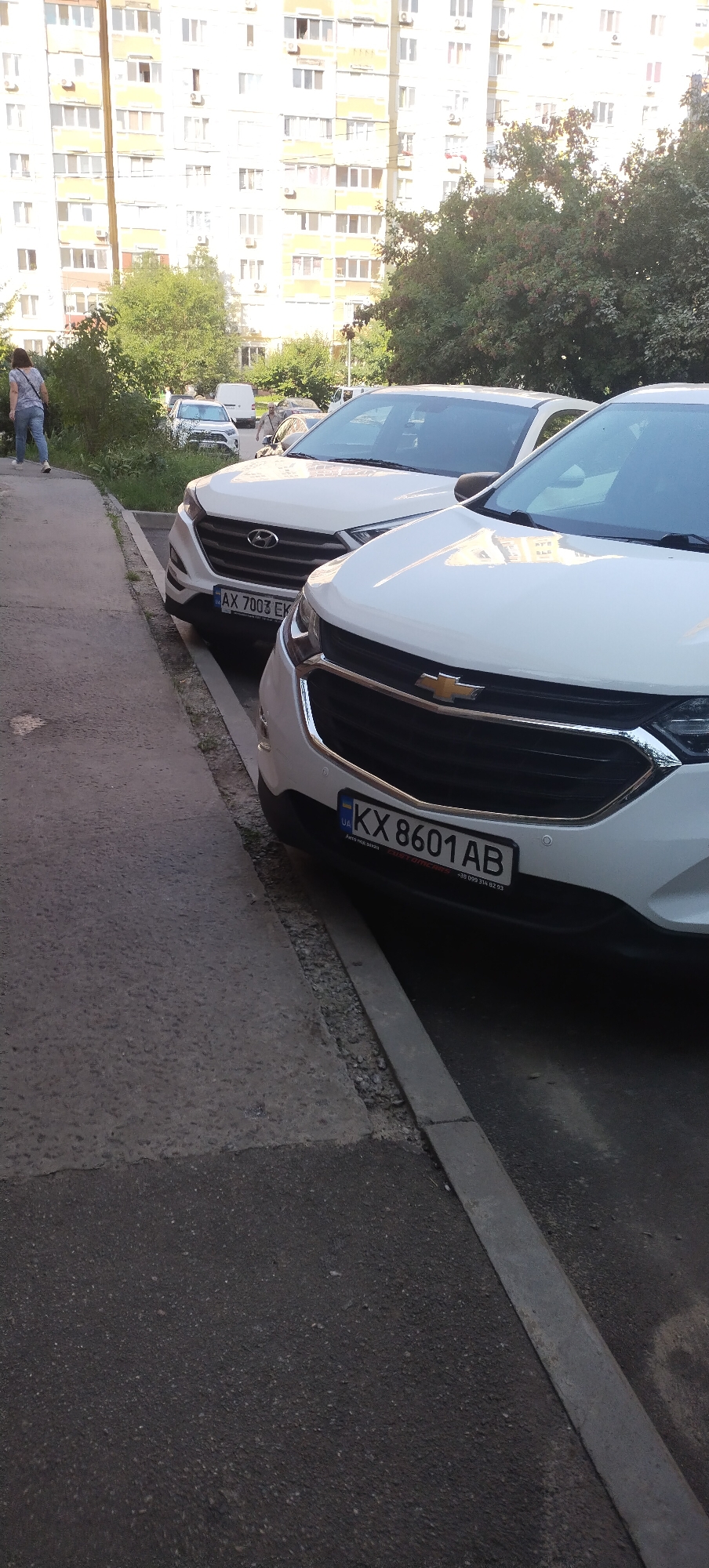 The image shows a street scene in Pisochyn, Ukraine, with two white cars parked on the side of the road. In the foreground, a white Chevrolet is parked, its license plate reading "KX 8601 AB". Behind it, a white Hyundai SUV is also parked, with the license plate "AX 7003 EK". A woman in jeans and a t-shirt is walking away from the camera on the sidewalk to the left of the cars. In the background, a multi-story building with numerous windows and balconies is visible, along with trees and other parked vehicles. The scene appears to be in a residential area, with a sidewalk and road.