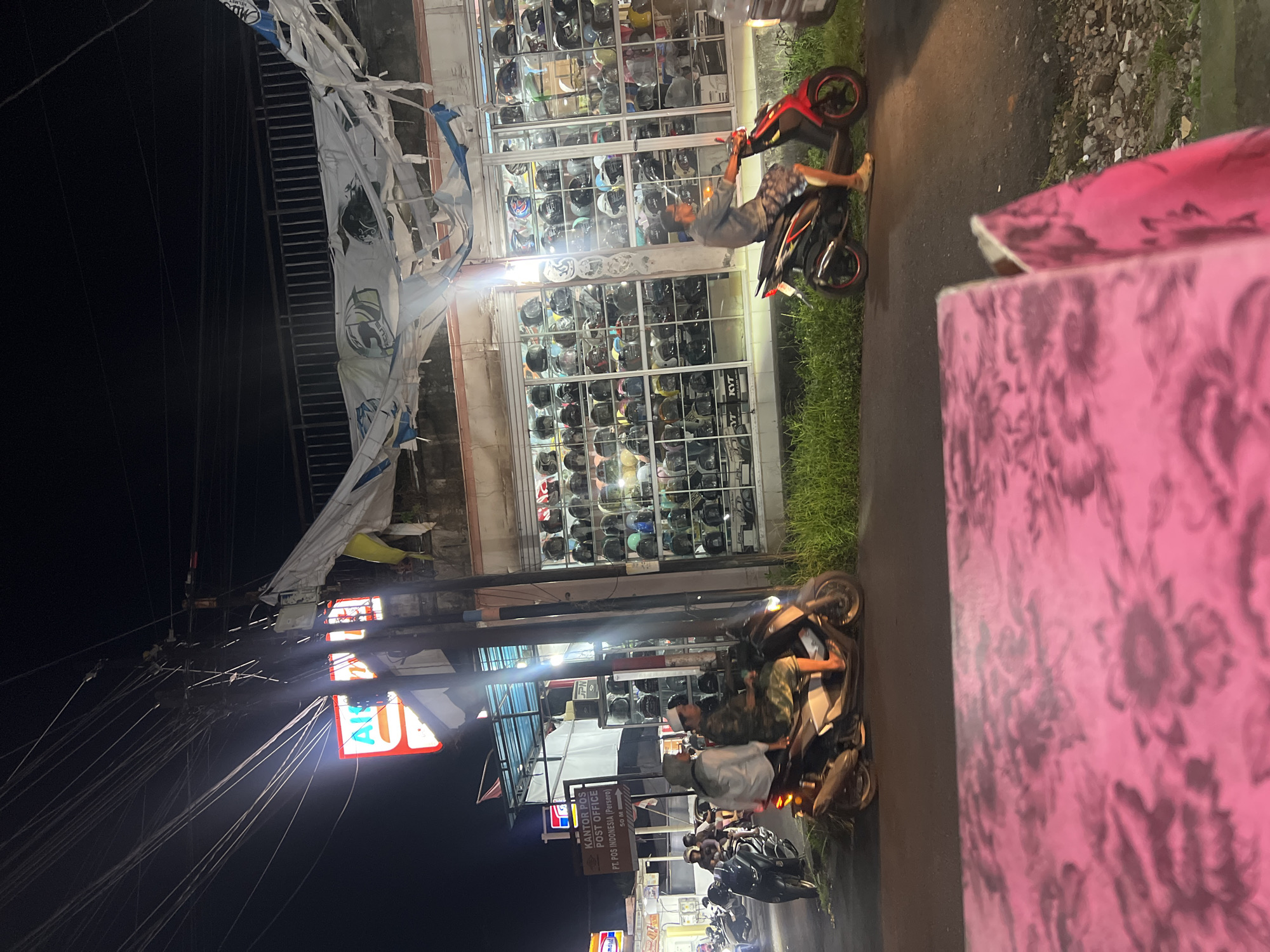 At night on a street in Gianyar, Indonesia, a store with a large display of helmets is illuminated. The store's facade features rows of shelves filled with various helmets behind a glass window. A torn banner hangs above the entrance. Two men are riding motorcycles on the street in front of the store. One man on a white and gray scooter is wearing camouflage pants and a cap. Another man on a red scooter is wearing a gray shirt and patterned shorts. In the background, a sign for "KANTOR POS POST OFFICE PT. POS INDONESIA (Persero)" is visible, indicating the presence of a post office. The street is paved, with grass and gravel on the side. Overhead, numerous electrical wires are strung between poles.