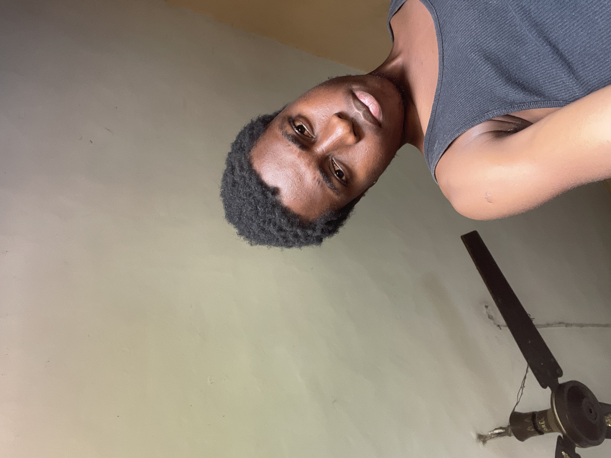 A medium-shot photograph, slightly tilted, features a dark-skinned man with short, dark hair and a black tank top. He is positioned towards the top of the frame, looking down and to the right of the camera. His face is well-lit, highlighting his eyes, nose, and mouth. The background is a pale, off-white wall with some slight imperfections. In the lower right corner of the image is a dark-colored ceiling fan. This image was likely taken indoors. No specific details link the image to Onitsha, Nigeria.