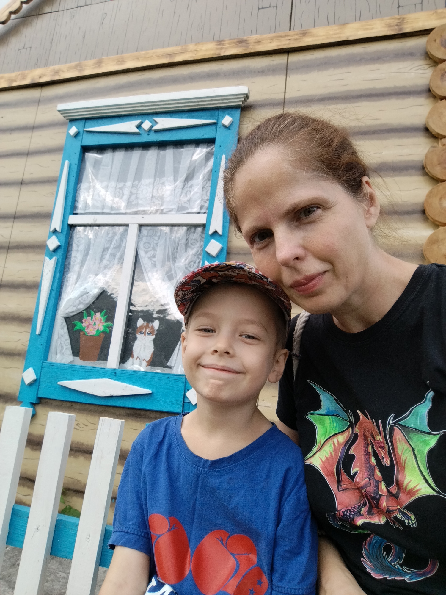 A medium shot shows a woman and a young boy taking a selfie outdoors, with a wooden building in the background. The building has a decorative blue window with white trim. The boy, wearing a blue t-shirt and a baseball cap, is in the foreground, smiling. The woman, wearing a black t-shirt with a dragon graphic, is to his right. The image was taken in Zavodoukovsk, Russia.