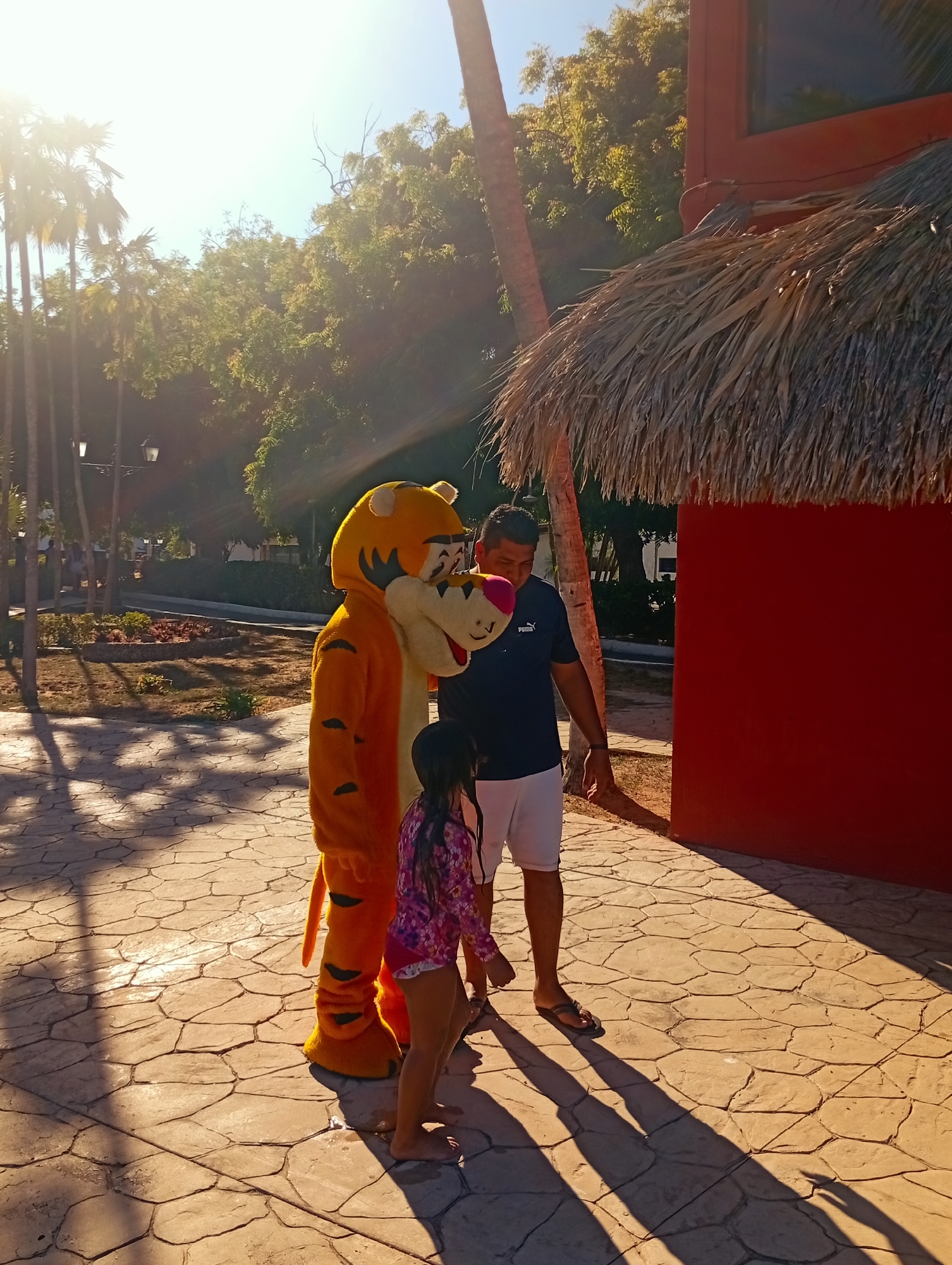 A man, a young girl, and a Tigger mascot stand on a paved area beside a red building with a thatched roof. The man wears a navy t-shirt, white shorts, and flip-flops. The girl wears a pink and purple floral long-sleeved shirt and swim bottoms. Tigger is orange and black. The paved area is textured with an irregular pattern. Palm trees and other trees are in the background. The sun is bright, casting long shadows. The location is Playa El Agua, Venezuela.