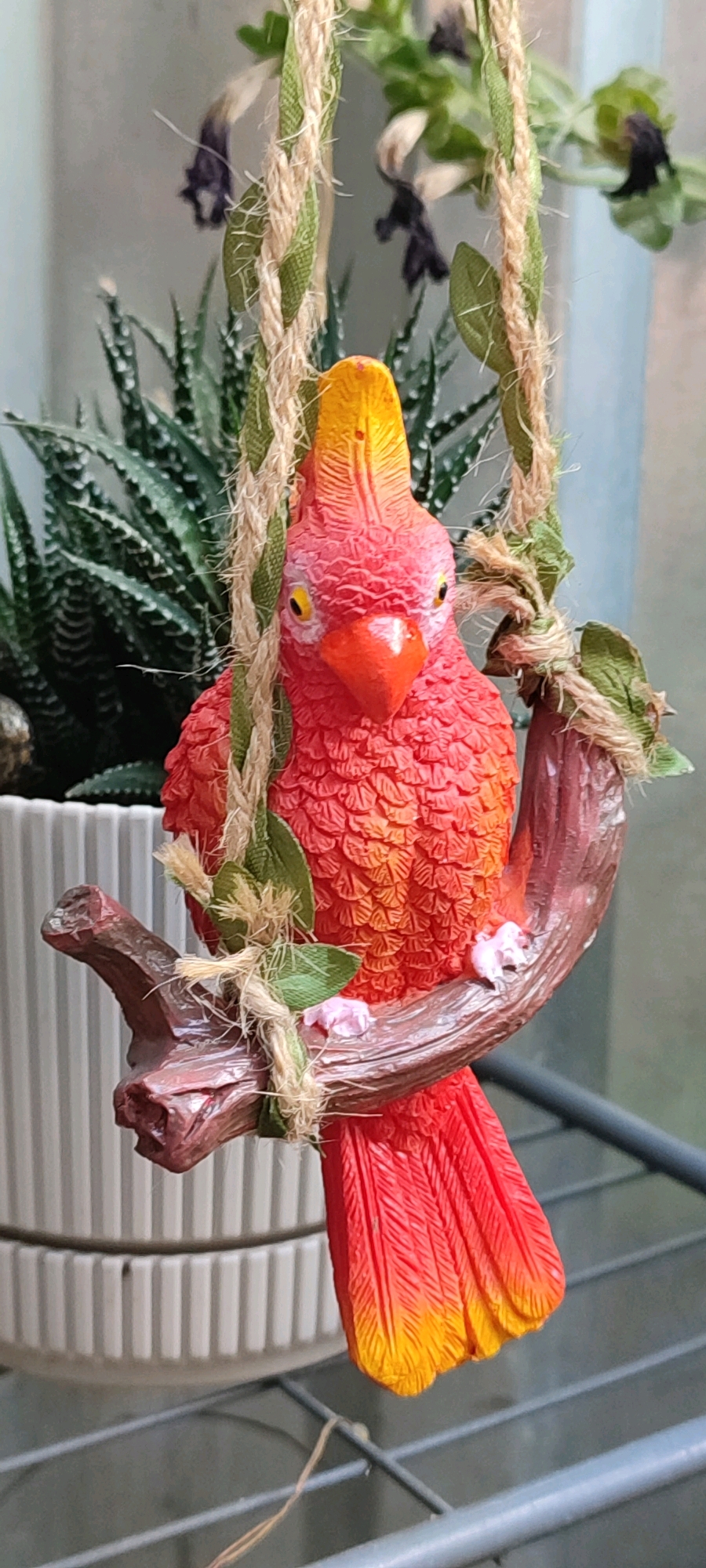 A close-up, eye-level shot features a decorative, brightly colored parrot figurine hanging from a jute rope adorned with artificial leaves. The parrot, predominantly red and orange with yellow accents on its crest and tail, is perched on a simulated branch. Tiny pink floral accents are visible on the branch, near the parrot's feet. In the background, out of focus, is a green, spiky succulent plant in a ribbed white pot. The overall setting appears to be indoors, possibly on a shelf, with a muted gray background suggesting a wall or distant structure. No specific location in Belaya Kalitva, Russia can be determined from the image.
