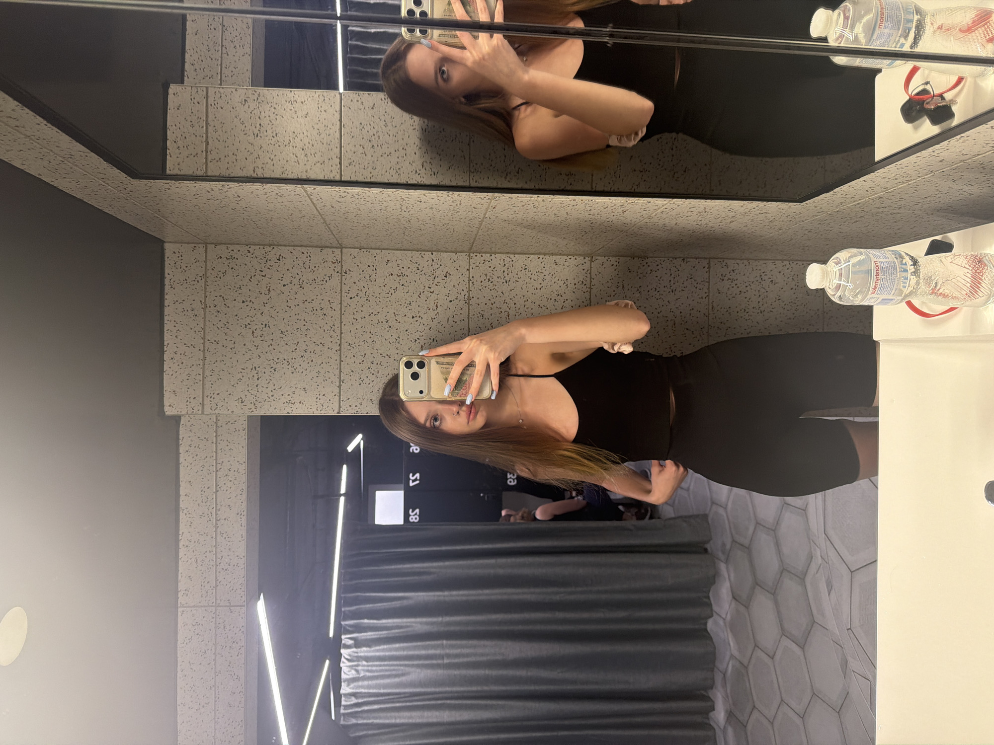 A young woman is taking a mirror selfie in what appears to be a public restroom or fitting room. She is wearing a black tank top and black shorts. Her hair is long and brown, and she has light-colored nail polish on. In her right hand, she is holding her phone, which has a light-colored case. She is looking at her reflection in the mirror, and her expression is neutral.
The setting includes a tiled wall with a speckled pattern, a large mirror, and a sink area. There are also some bottles of water and keys visible on a shelf in the mirror's reflection. The lighting is bright and appears to be artificial, suggesting an indoor environment. The overall mood is casual and personal, as indicated by the selfie. There is some text visible on the water bottle label, but it is not legible in the image.
