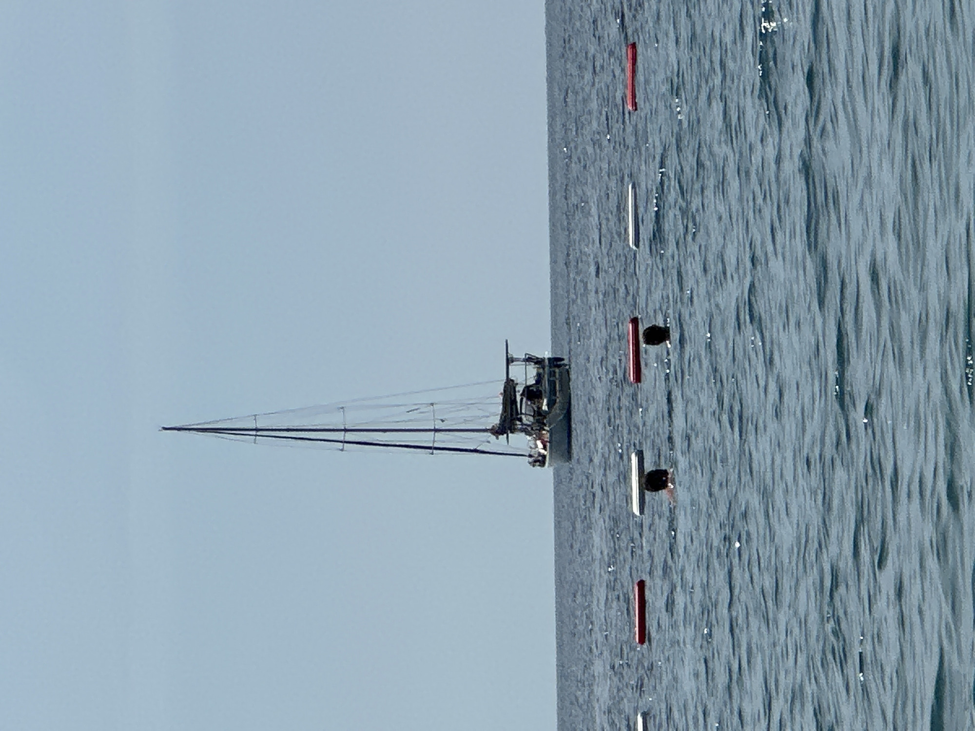 A sailboat is sailing on the water, marked by several red and white buoys. The sky is clear and light blue. The water is textured with small waves. The location is not discernible from the image.