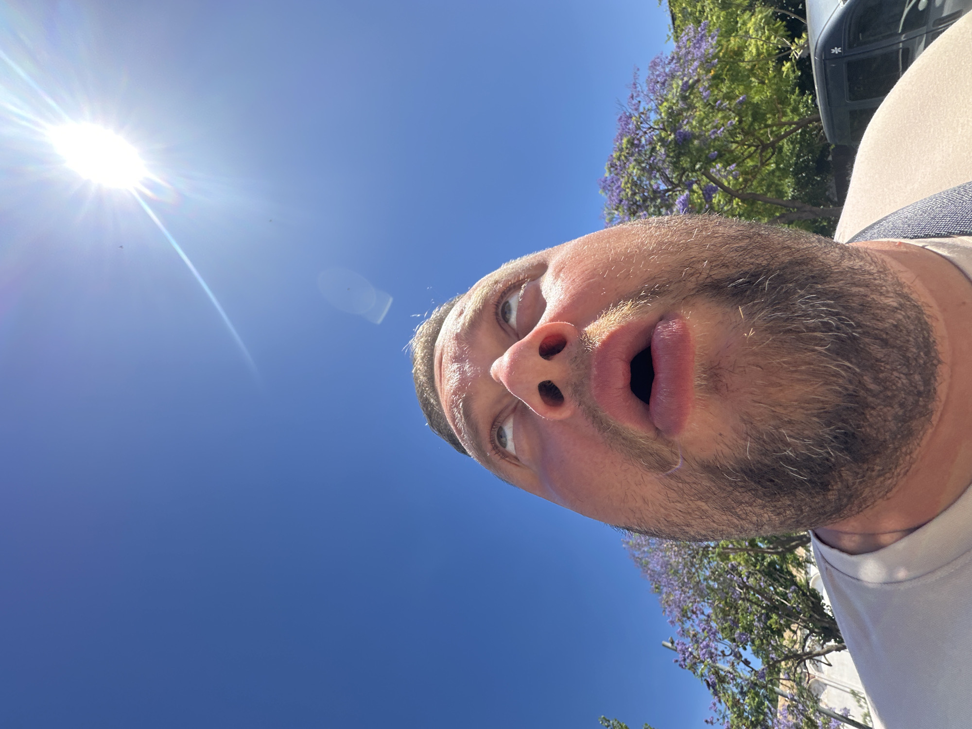 This is a low-angle, close-up, outdoor, eye-level shot of a man's face and upper chest. The man is looking upwards, with his mouth open in a surprised or awestruck expression. His eyes are wide, and his eyebrows are raised. He has short, dark hair and a beard. The sun is shining brightly in the upper right corner of the frame, creating a lens flare and casting a bright light on his face. In the background, there are trees with purple flowers, likely jacaranda trees, and a clear blue sky. The man is wearing a light-colored t-shirt and a strap across his shoulder. A black van is partially visible on the right side of the frame. The scene appears to be set in Sant Feliu de Guíxols, Spain, given the presence of jacaranda trees which are common in the region during spring and summer, and the architectural style visible in the background hints at Mediterranean influences.