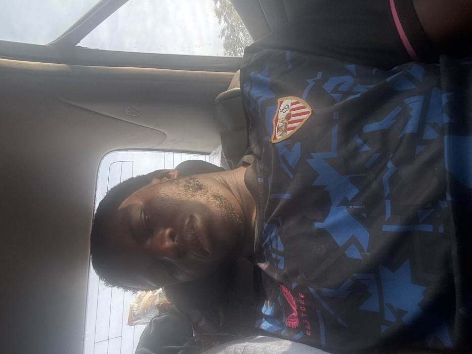 A man is reclined in a car seat, appearing drowsy or asleep. He is wearing a dark sports jersey with a prominent crest or logo on the chest and blue geometric patterns. The word "CASTORE" is visible on the shirt. The car's interior includes a sunroof or window above. The lighting is somewhat harsh, with some areas overexposed.