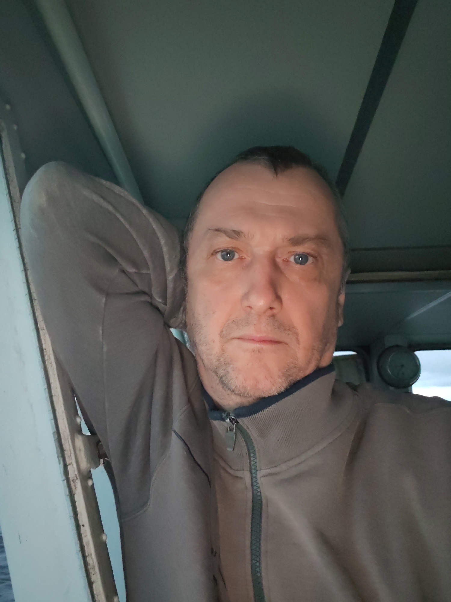 A medium shot of a middle-aged man with light skin and blue eyes. He has short brown hair and a light beard. He is wearing a brown zip-up jacket and is leaning against a white metal structure with his arm raised. The background appears to be a part of the interior of a vehicle or structure with light blue walls and ceiling. A round gauge is visible in the background. The location is Unknown, Unknown.