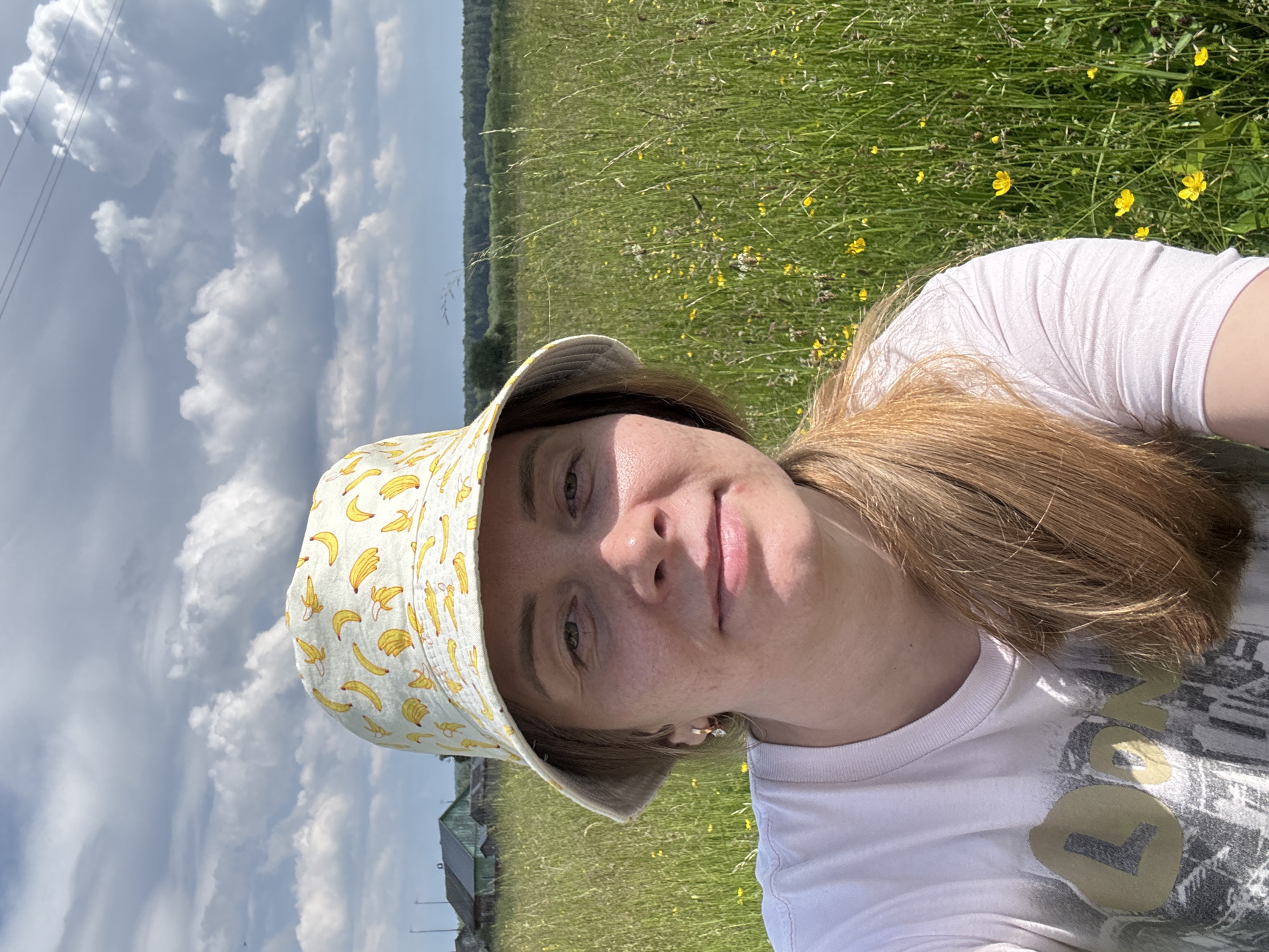 A woman is outdoors in a field with tall green grass and scattered yellow flowers. She is wearing a white bucket hat with a banana print and a light pink t-shirt with a graphic design. Her light brown hair is pulled back and falls over her shoulder. In the background, there are some rustic buildings and a treeline. The sky is partly cloudy. The image appears to have been taken in a rural area, possibly on the outskirts of the city of Kurgan, Russia, given the presence of traditional wooden houses and open fields.