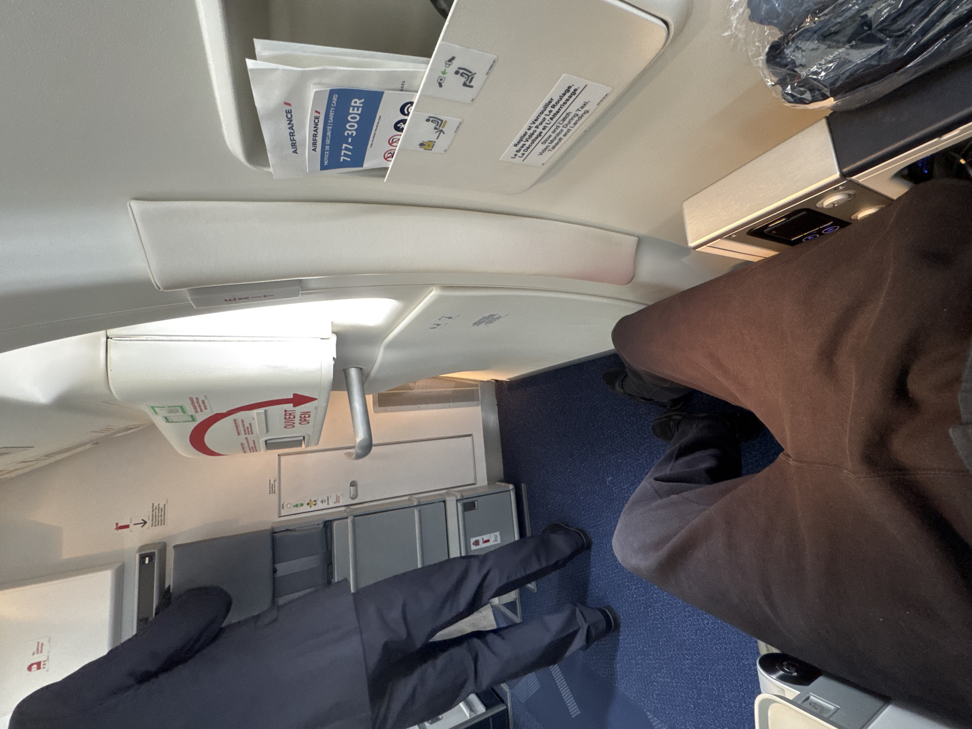 This is an interior shot of an Air France 777-300ER aircraft, likely taken while in flight, potentially taxiing, or during boarding. Visible overhead are the passenger information and emergency exit indicators, with a safety card in the storage compartment labeled "Air France/Notice de Sécurité | Safety Card 777-300ER". The image also shows the overhead reading light and ventilation controls. The emergency exit sign is marked with "OUVERT" and "OPEN" as well as a red arrow, with directions of operation and pictorial instructions.
The blue carpeted aisle is partially visible. Two sets of human legs are present, one dressed in what appears to be a dark suit or similar attire, and the other in brown sweatpants, in the foreground. The location appears to be inside an aircraft, and not in Le Mesnil-Amelot, France.
