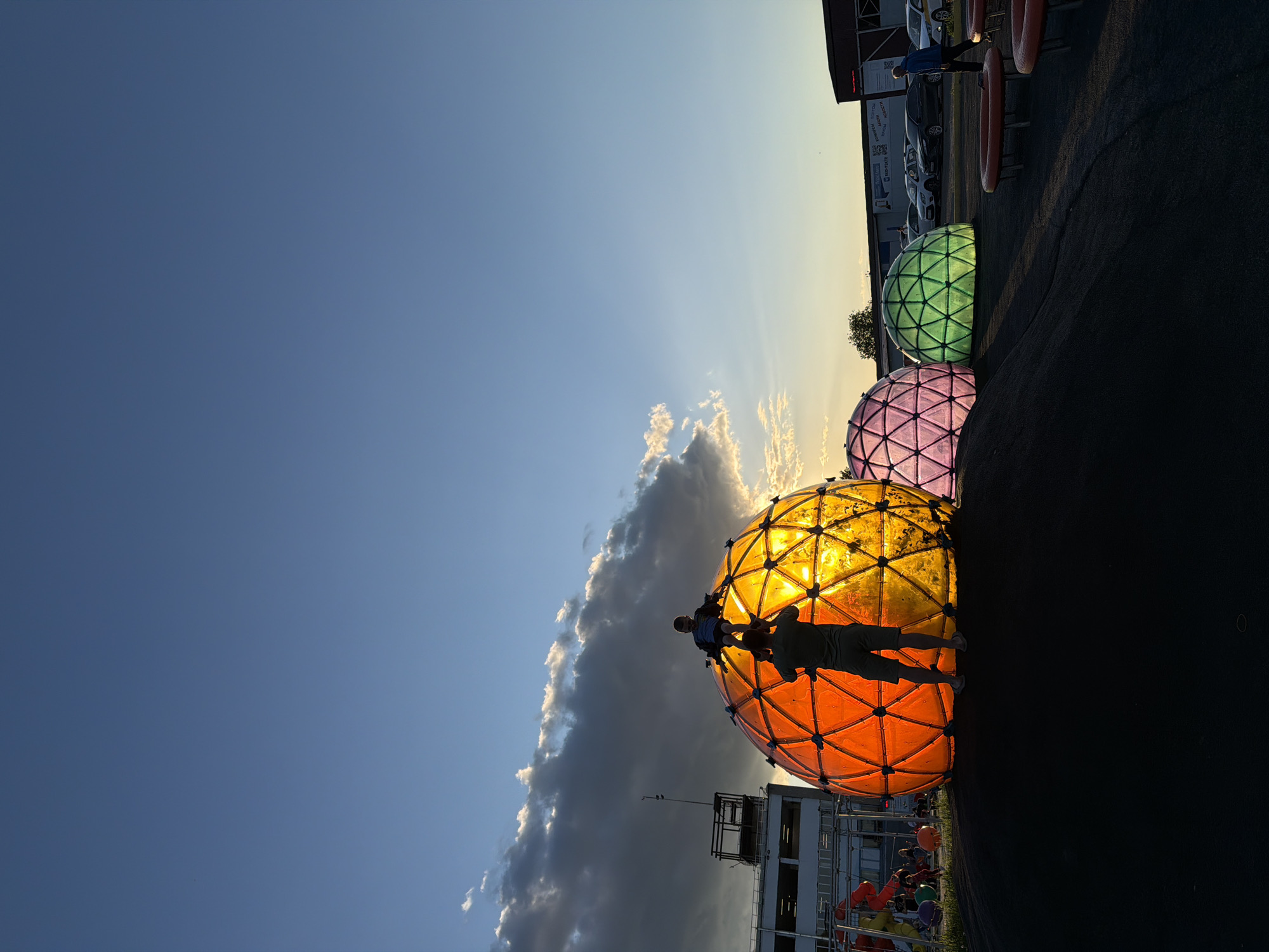 A low-angle, wide-shot captures three geodesic spheres illuminated in orange, pink, and green hues. Two silhouetted people are standing on the orange sphere. The sky is partly cloudy, with blue visible in the upper-left and a yellow-orange glow from the sun at the top. The structures are located on an asphalt surface. A building is visible in the lower-center. Further behind the spheres, a building and several cars are parked. The location may be near Sin'kovo, Russia.