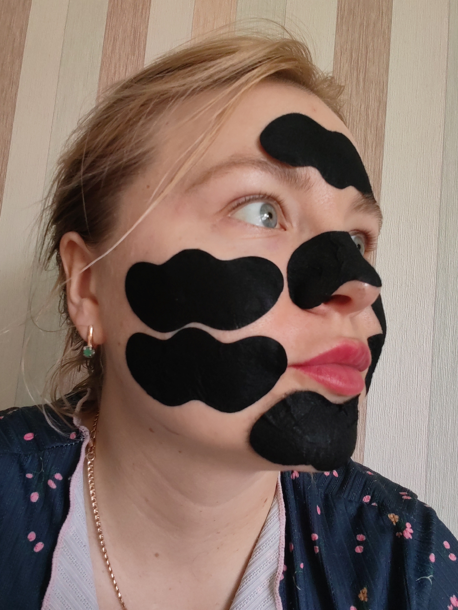 A fair-skinned woman with blonde hair is pictured indoors. She is wearing a blue and pink floral shirt, a gold chain necklace, and gold hoop earrings with green stones. Black facial strips are adhered to her forehead, nose, and cheeks. Her lips are pink. The background is a wall with beige vertical stripes. The location is in the city of Izhevsk, Russia.